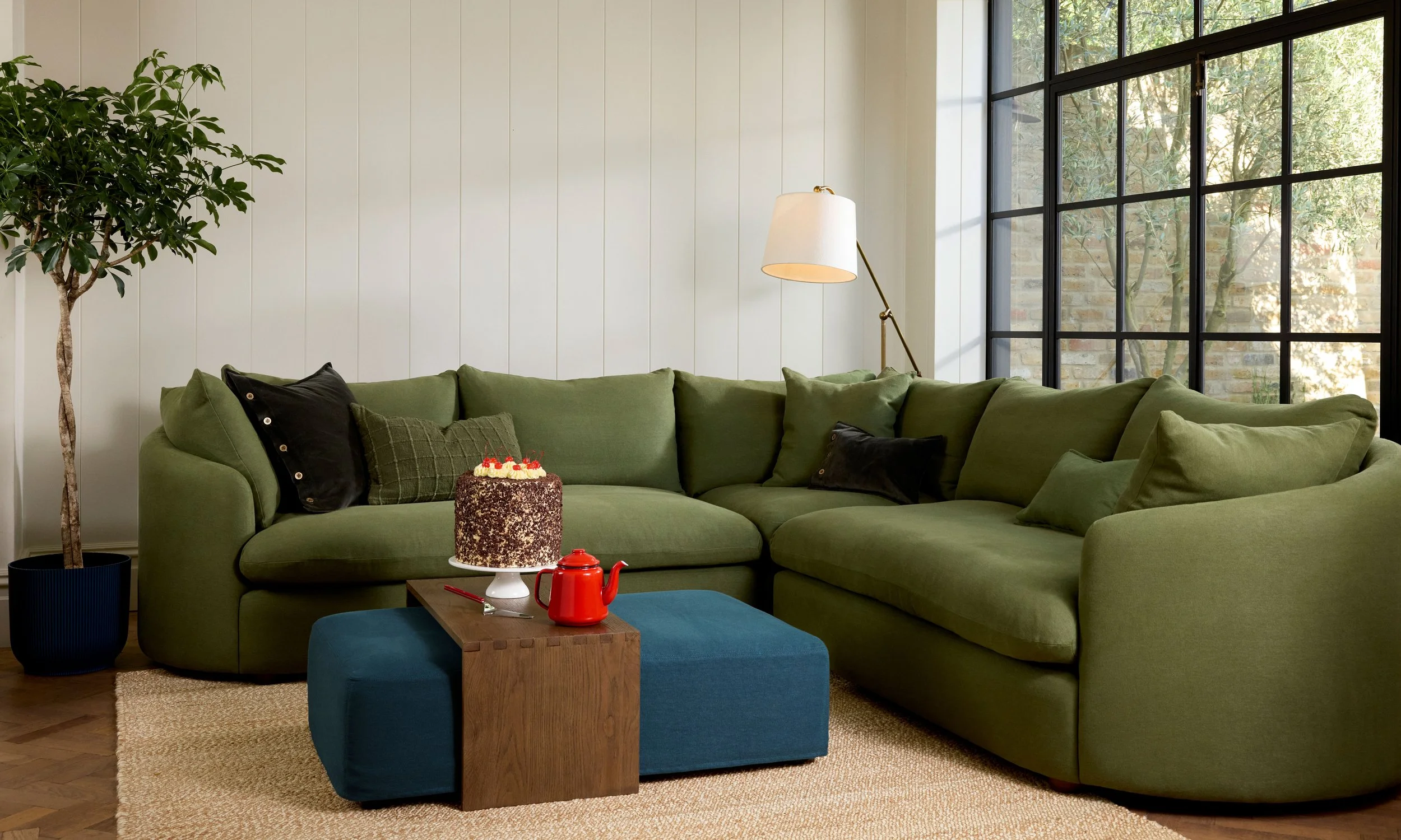 Living room with a large green sectional sofa, potted plant, floor lamp, window with black grid panes, and a blue ottoman with a birthday cake and red teapot on it.