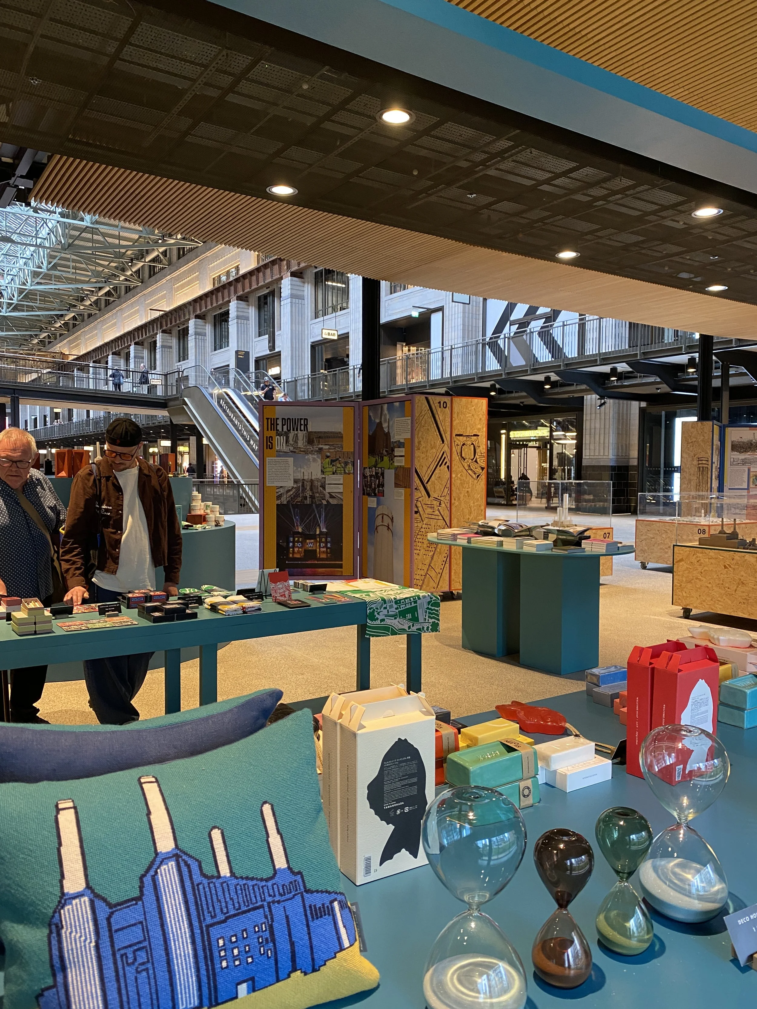 An indoor market or exhibition space with display tables selling various items, including books, packaged goods, and decorative objects. There are a few people browsing, and the background features a large open space with high ceilings, escalators, a
