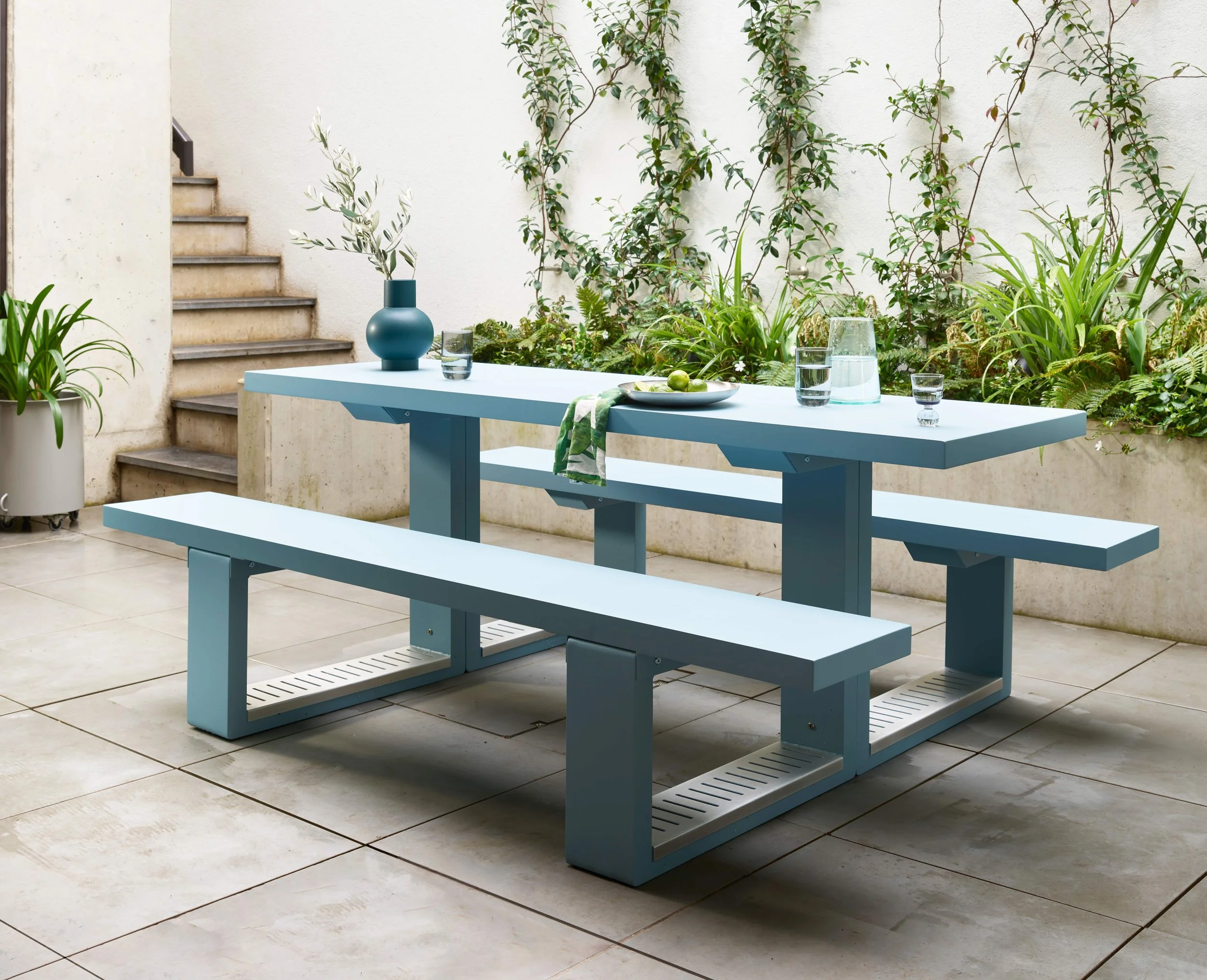 Light blue outdoor dining table with matching benches on a tiled patio, decorated with a blue vase holding green leaves, a plate with limes, glasses of water, and a green and white cloth; surrounded by greenery and stairs in the background.