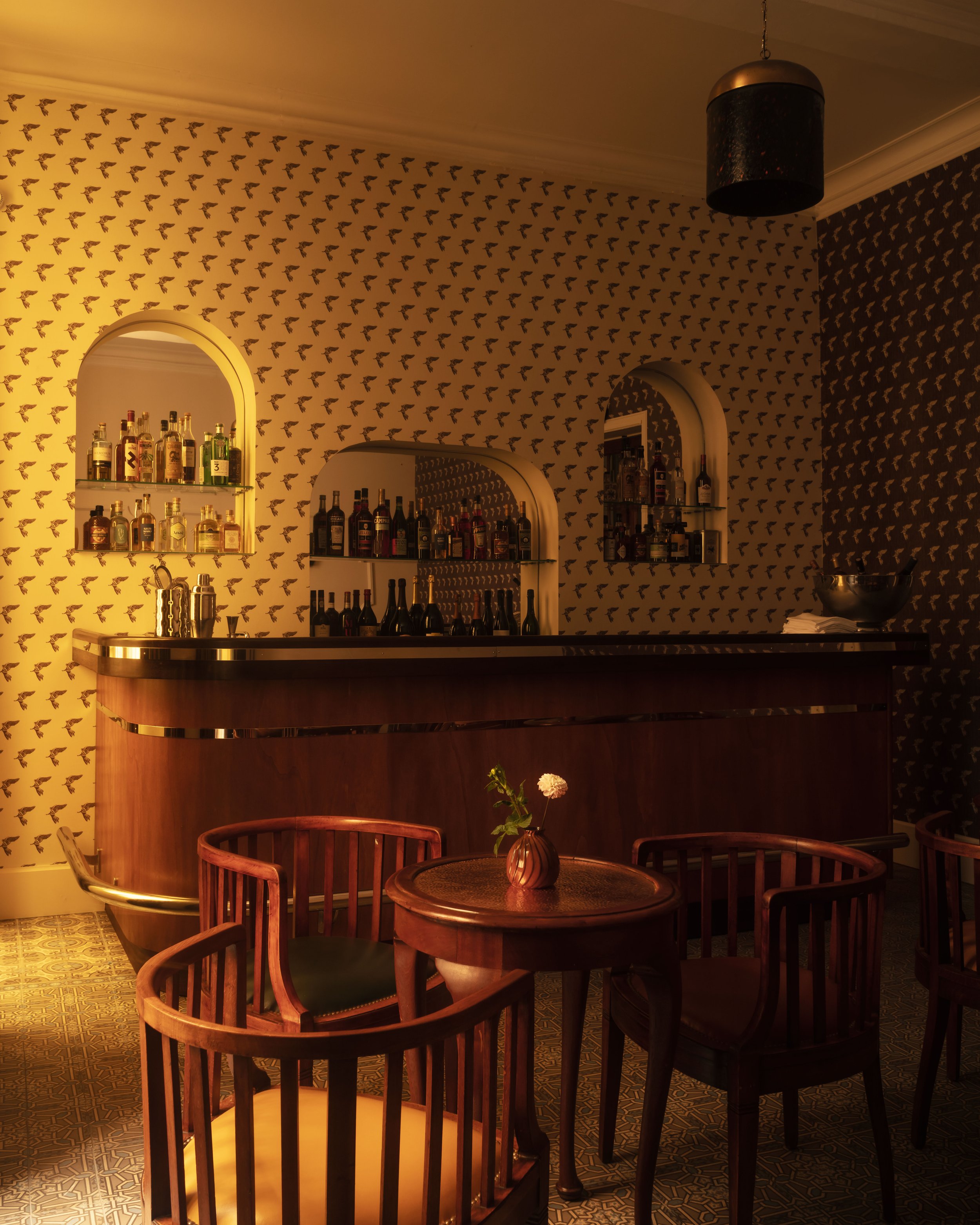 A dimly lit bar with a curved wooden counter, shelves of liquor bottles, a small round table with a flower in a vase, and patterned wallpaper with small bird designs.
