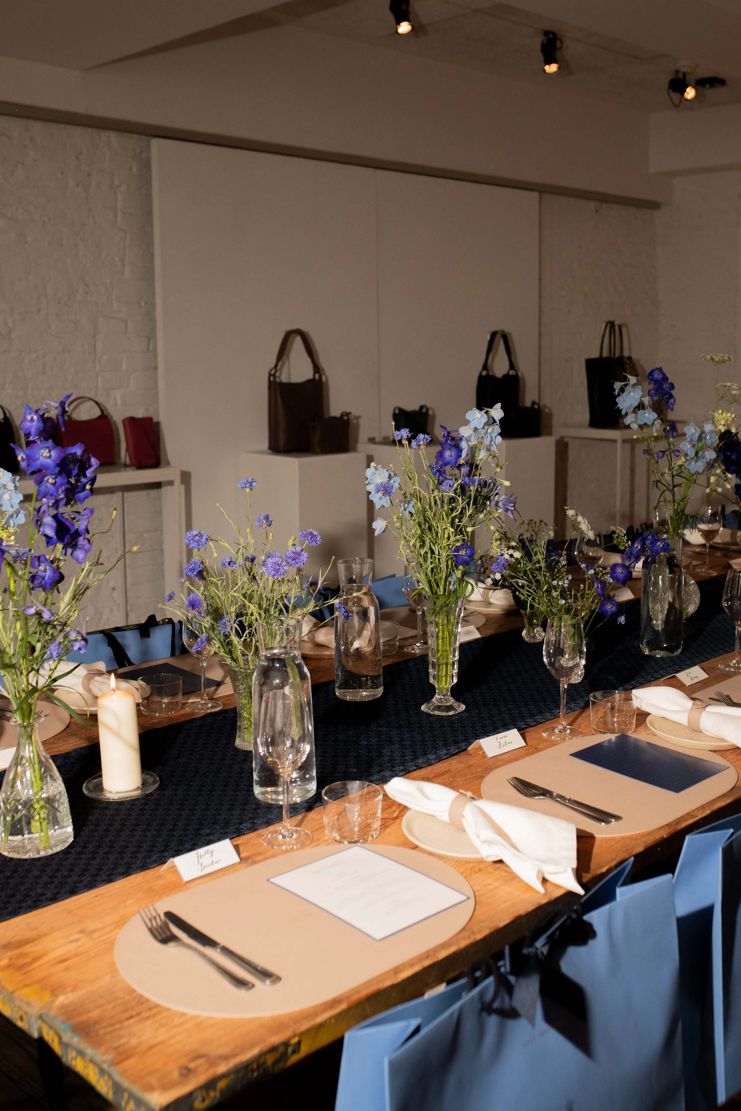 A decorated dining table with blue and purple flowers in vases, water pitchers, wine glasses, place settings with white napkins, candles, and gift bags along the side, in a room with white brick walls and handbags on display in the background.