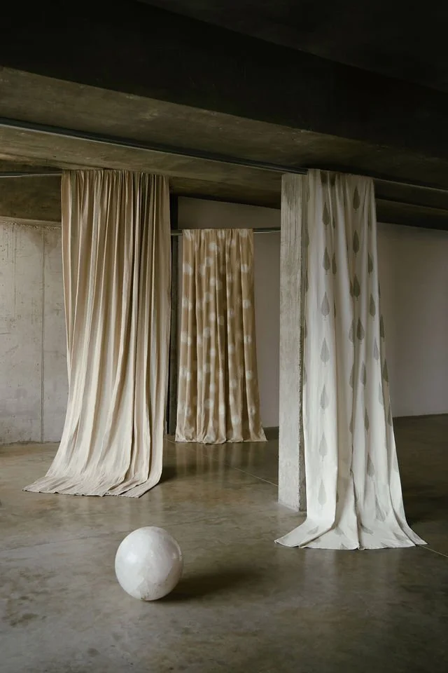 Three beige curtains hanging from the ceiling, with a white marble sphere on the floor in front of them, in an industrial-style concrete space.