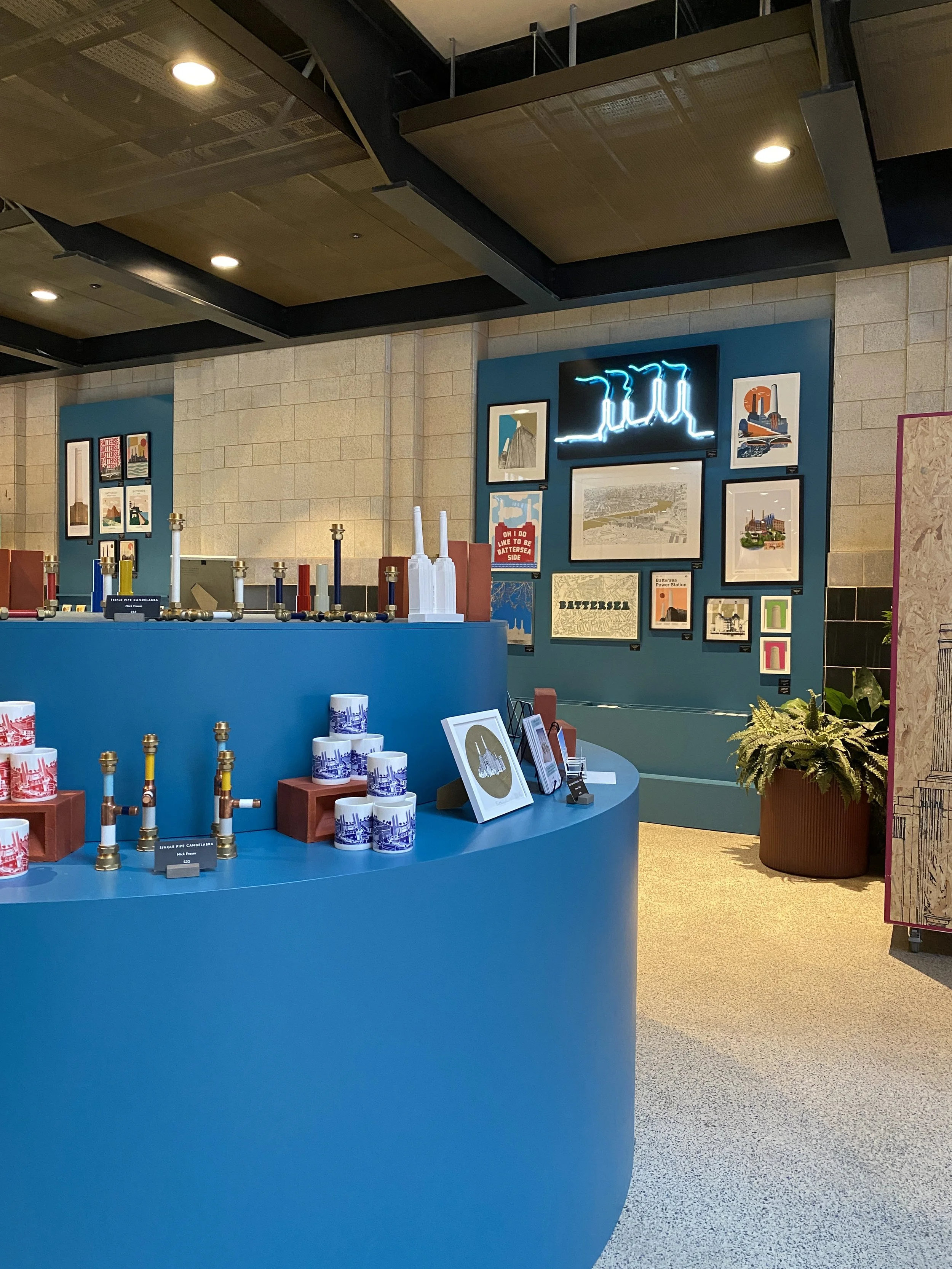 Interior of a modern space with a blue circular display table featuring decorative pipes, framed artwork on the wall, a neon sign, and a large potted plant.