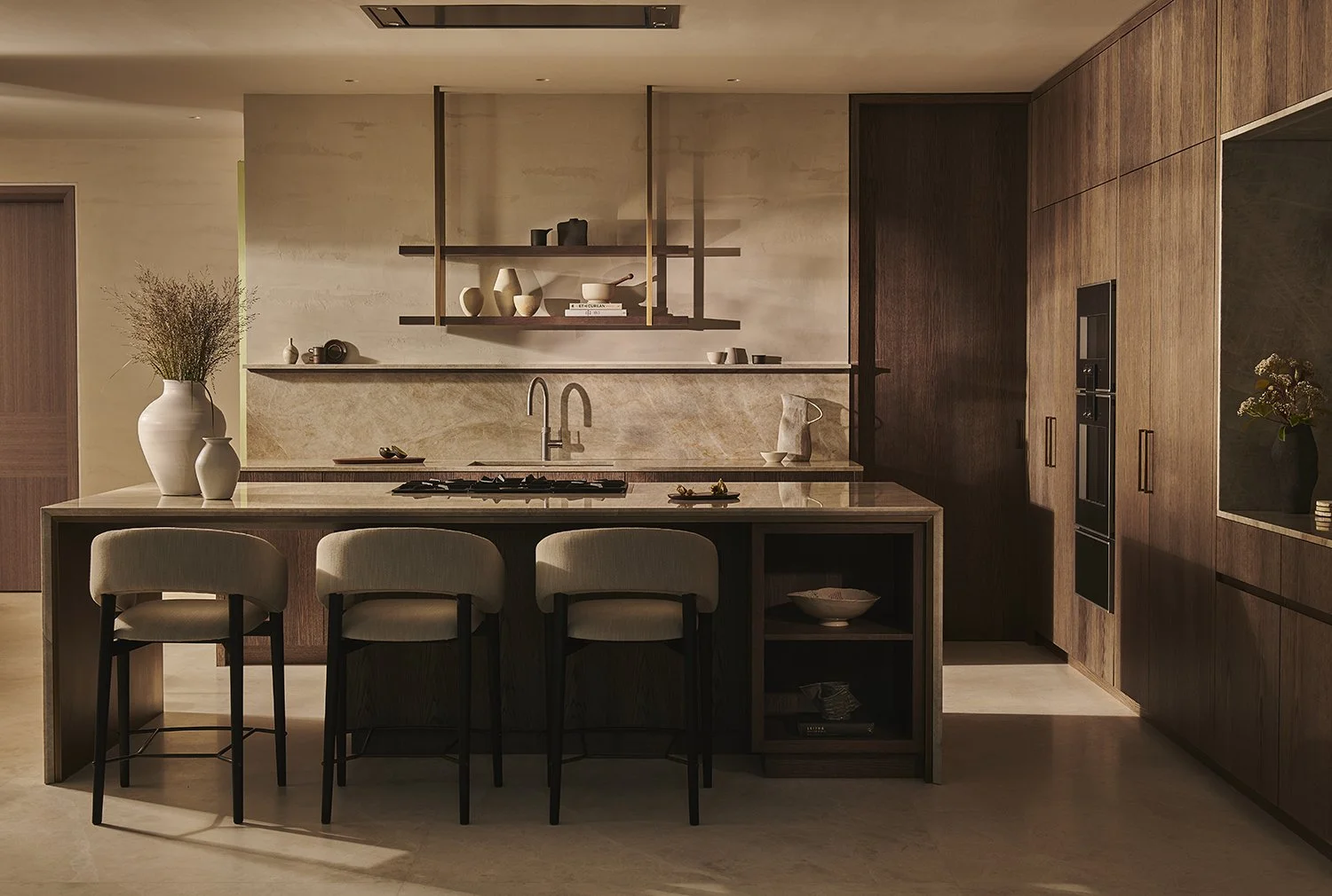 A modern kitchen with dark wood cabinets, a kitchen island with three beige chairs, open shelves above the counter with vases and books, a vase with dried flowers on the island, and a built-in oven and oven microwave on the right wall.