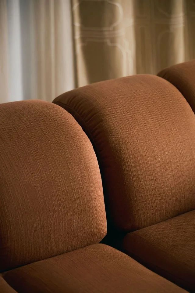 Close-up of a brown upholstered sofa with rounded armrests, in a warmly lit room with curtains in the background.