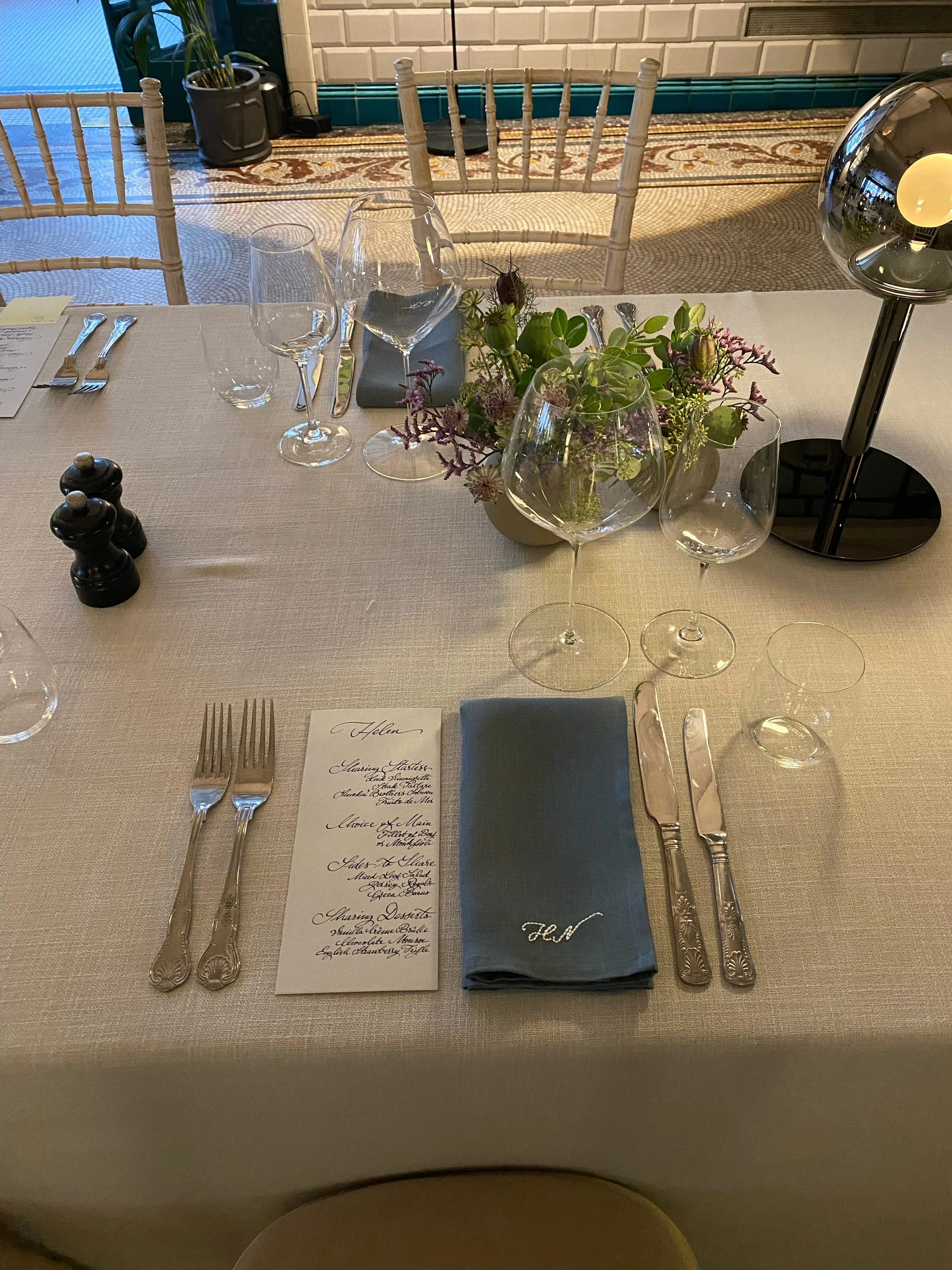 Elegant table set for a formal dinner with wine glasses, silverware, a menu card, a blue napkin with initials, a flower centerpiece, and a lamp on a white tablecloth.