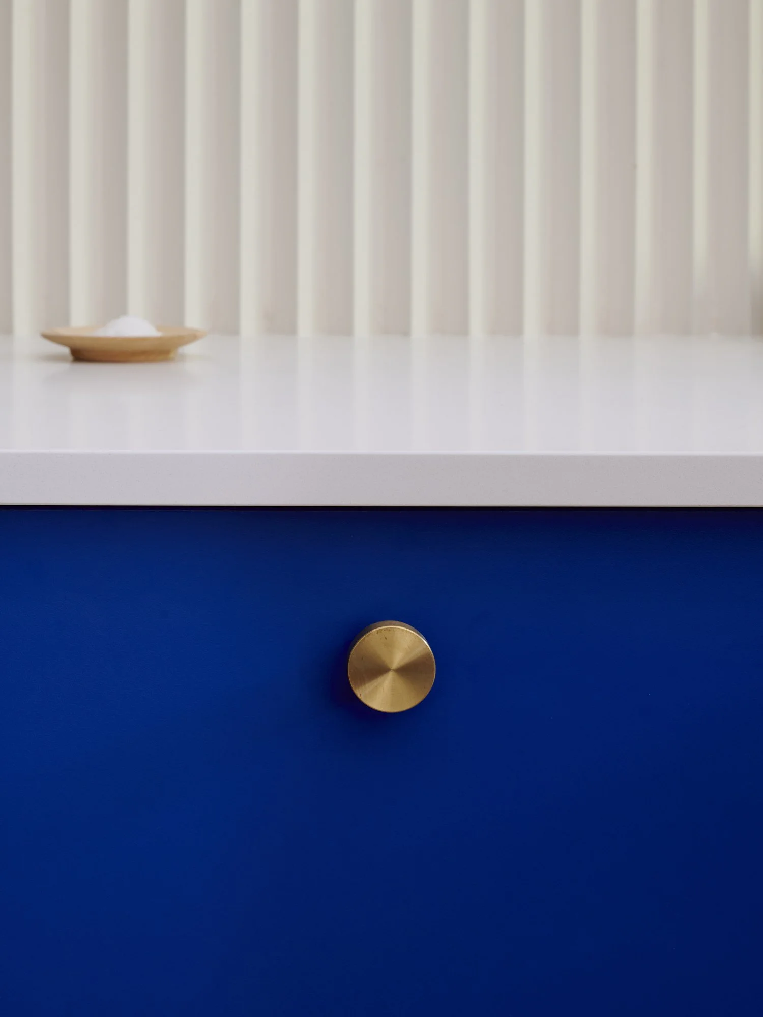 Close-up of a blue cabinet with a gold round knob and a white countertop with a small wooden dish containing white salt or sugar, set against a background with vertical white slats.