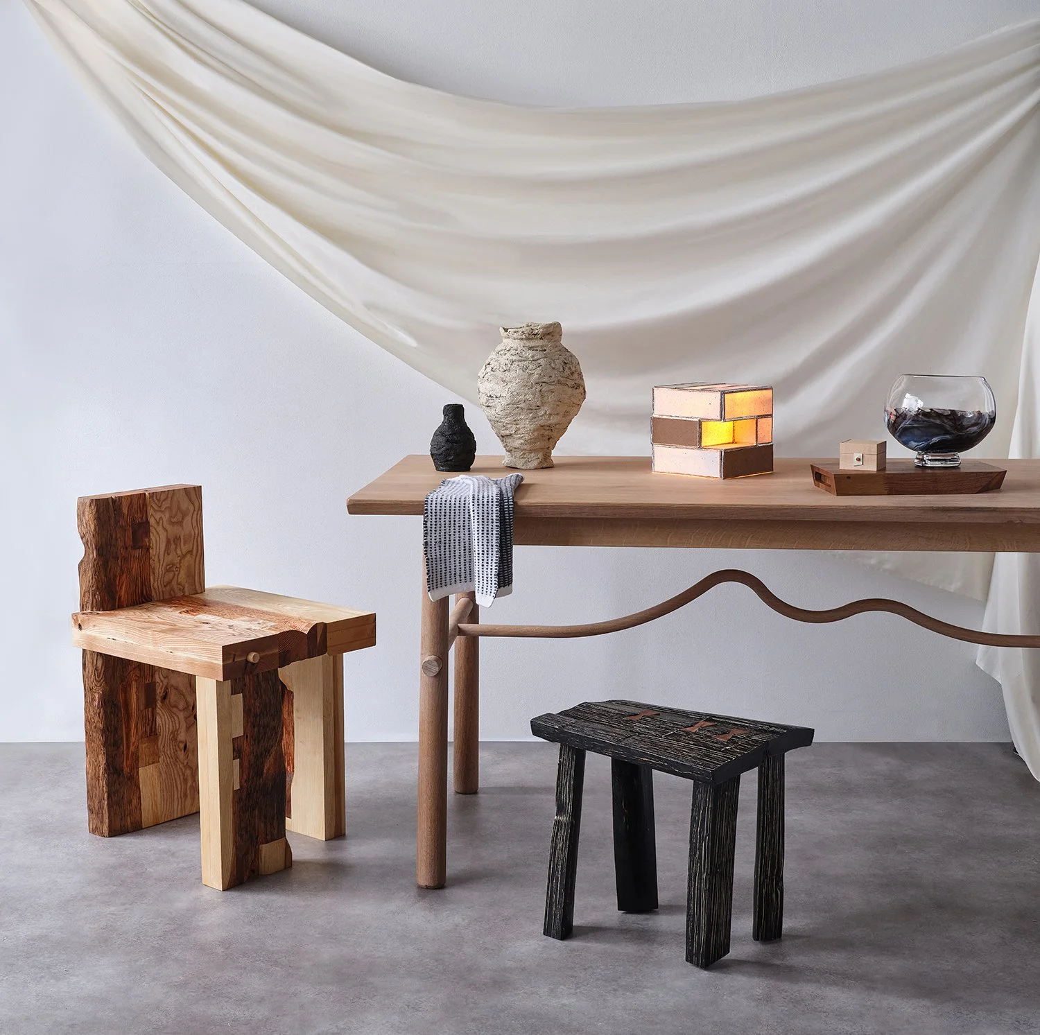 Minimalist room with wooden furniture, including a table and three stools, decorated with vases, a lamp, and a bowl of wine, against a backdrop of draped fabric.