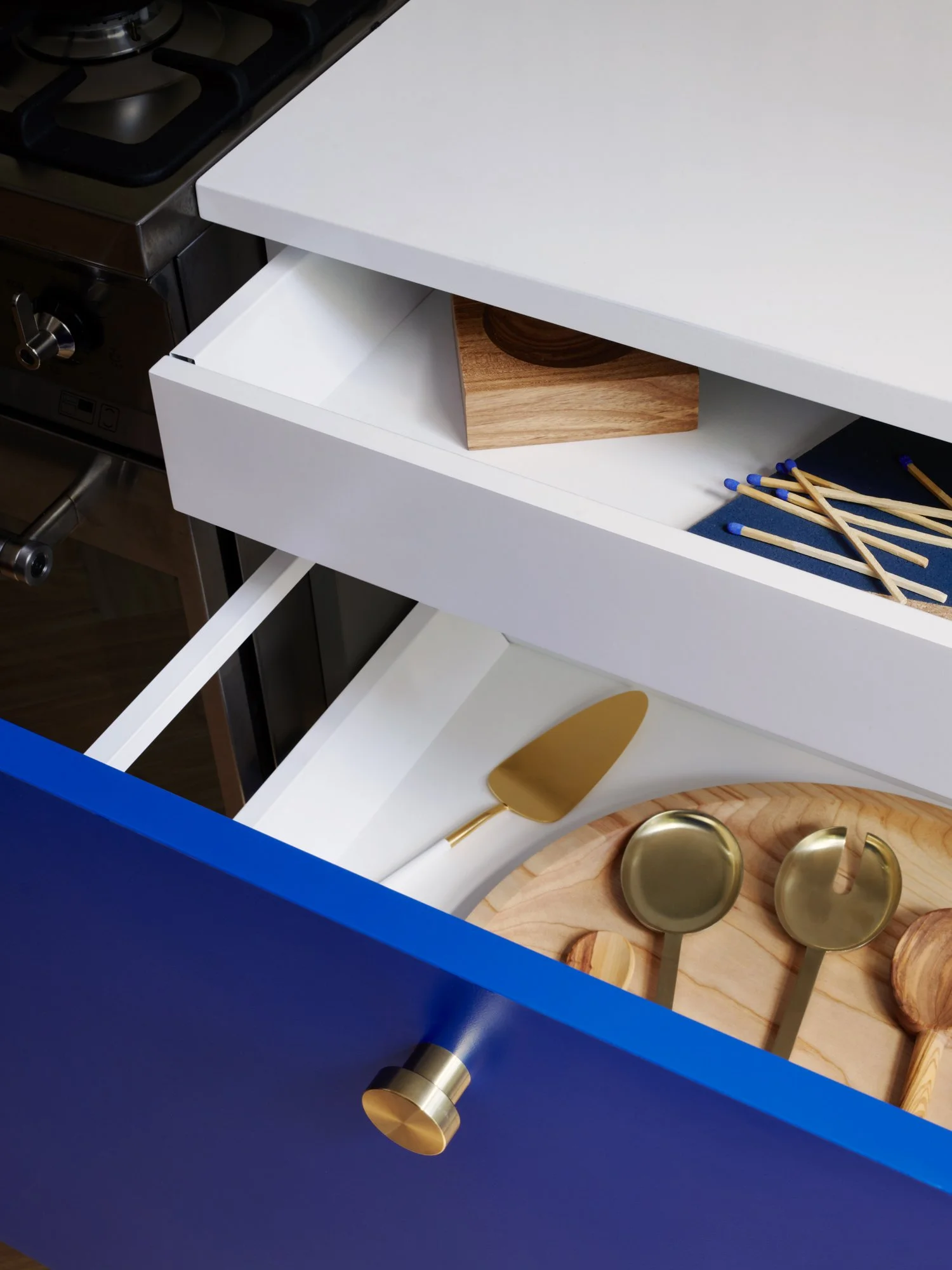 Open kitchen drawer containing gold kitchen utensils, including a serving spoon, on a wooden cutting board. The drawer is part of a kitchen cabinet with a blue exterior and gold knob. Extra utensils and a wooden block are visible in the background.