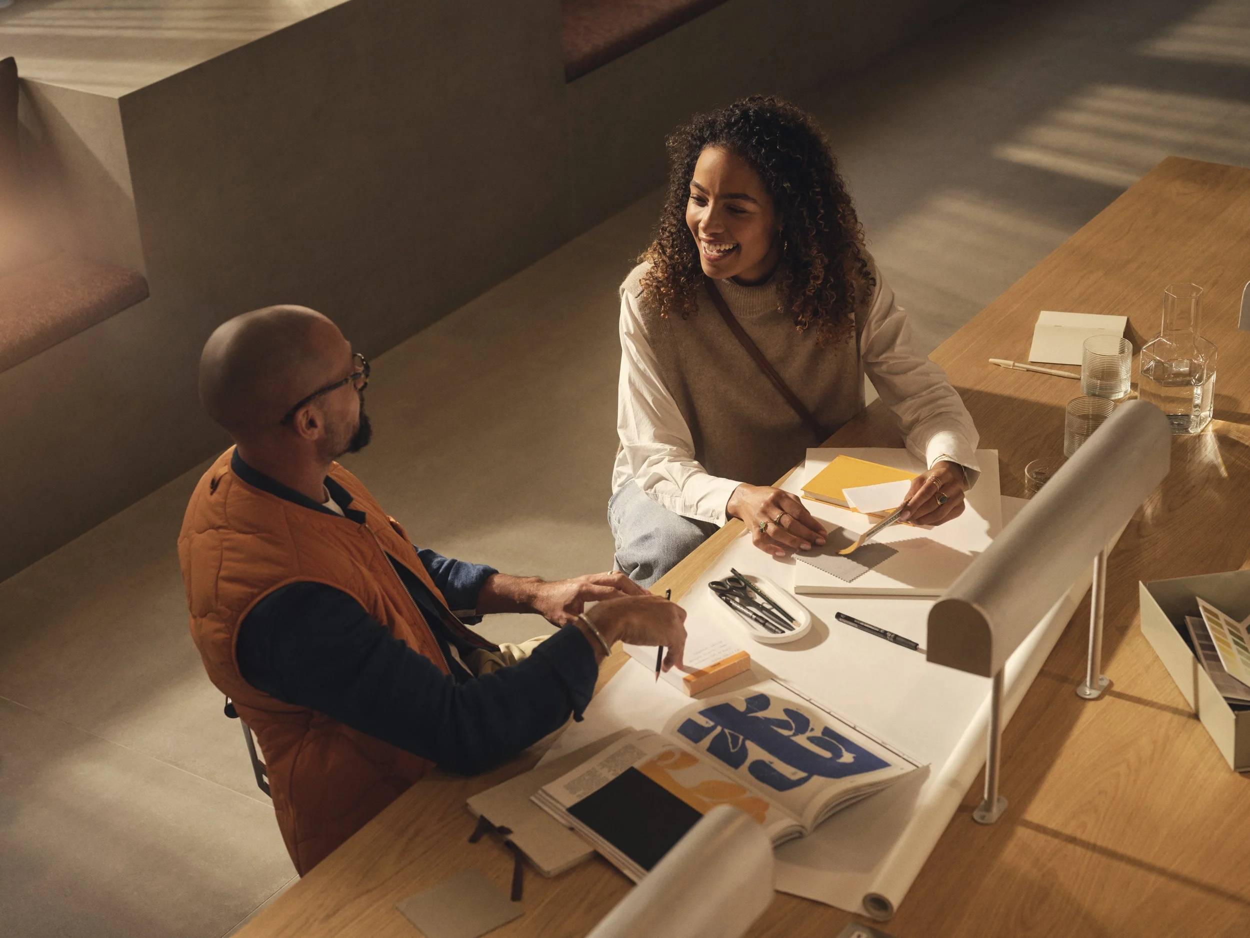 Two people sitting at a table, engaging in a lively conversation, surrounded by notebooks, papers, and art supplies in a well-lit, modern space.