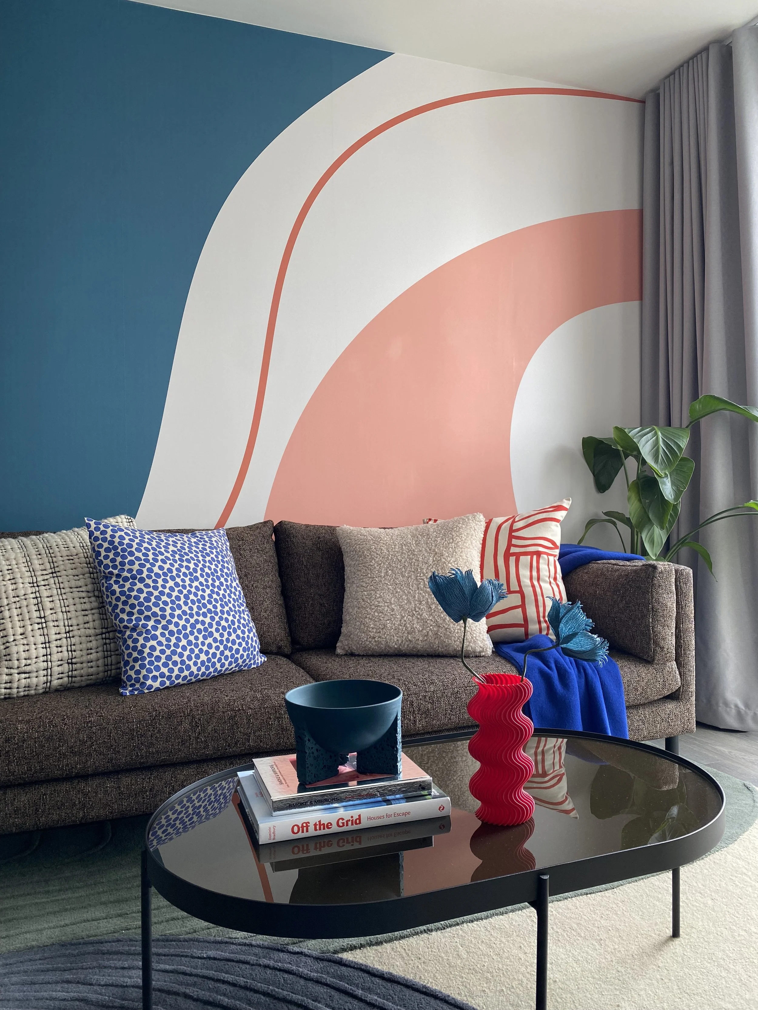 Modern living room with colorful abstract wall mural, brown sofa with patterned and solid cushions, a black coffee table with a red vase and books, and a potted green plant near gray curtains.