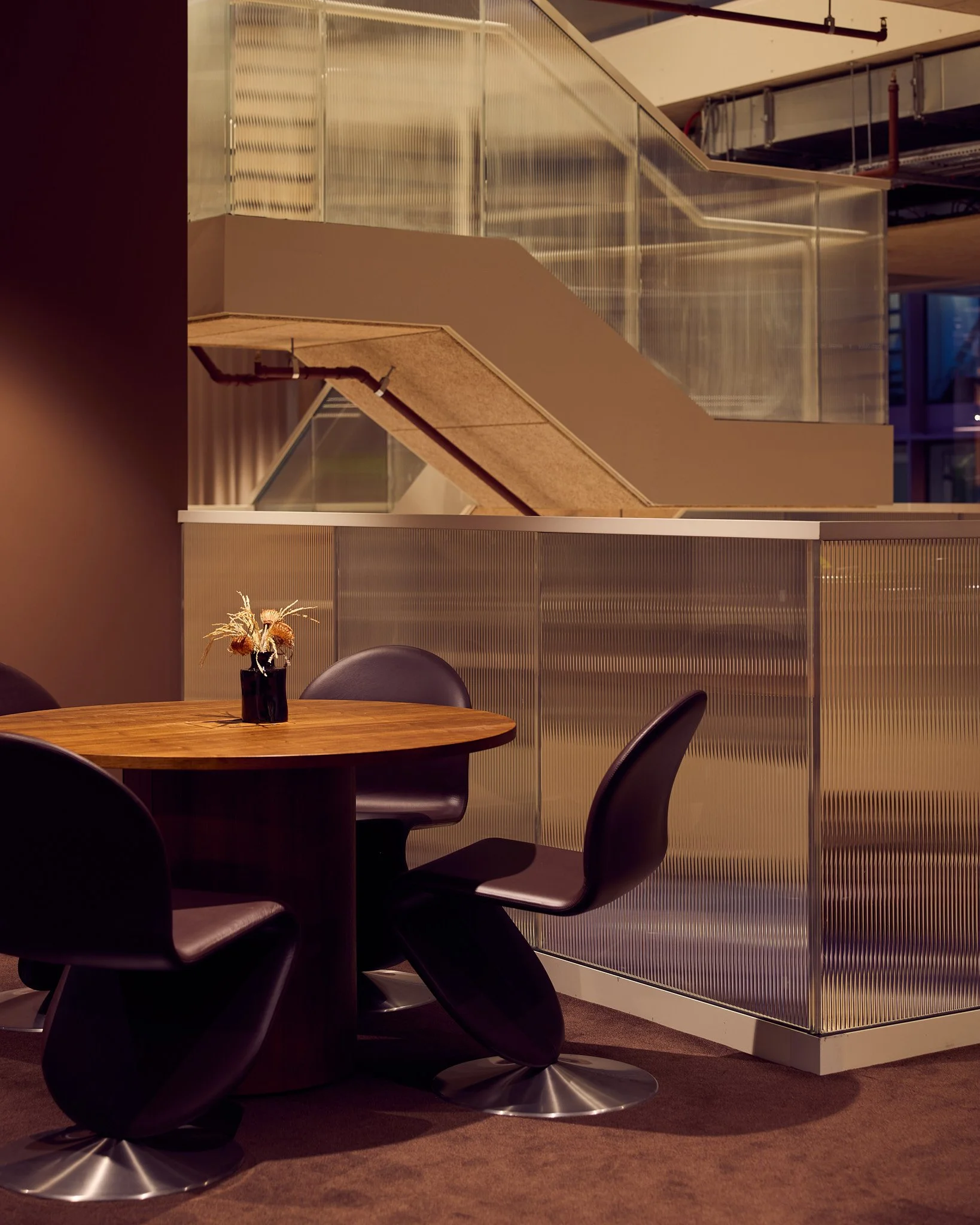 A round wooden table with three black chairs and a small floral centerpiece inside a modern interior space.