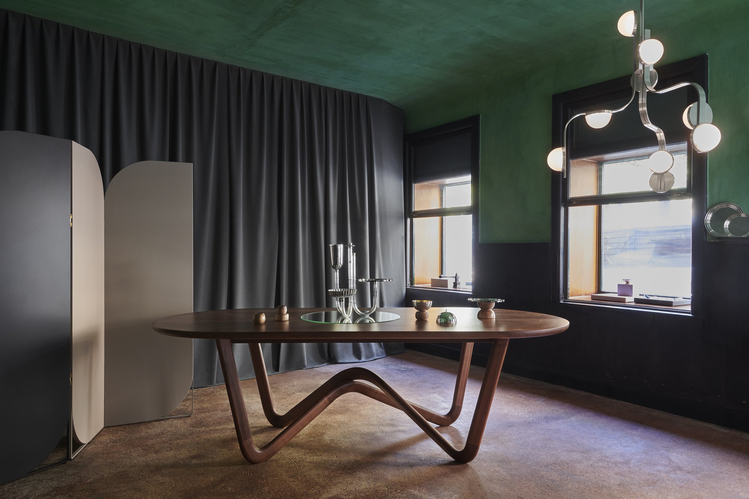 Interior of a room with a wooden oval table, decorative objects, black curtains, and large windows with natural light, featuring a green ceiling and a modern chandelier.