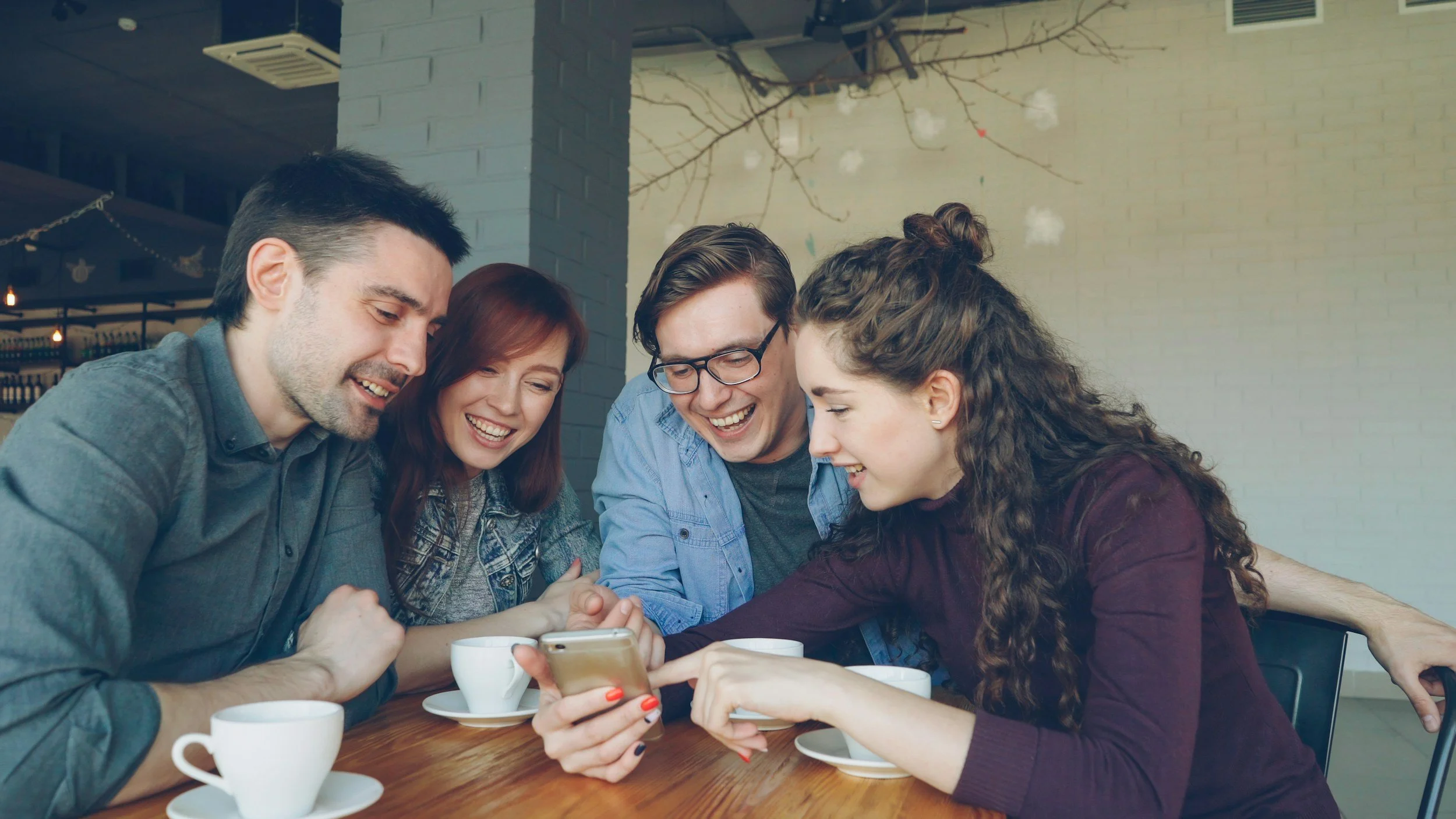 Five friends sitting at a table in a cafe, looking at a phone and laughing together.