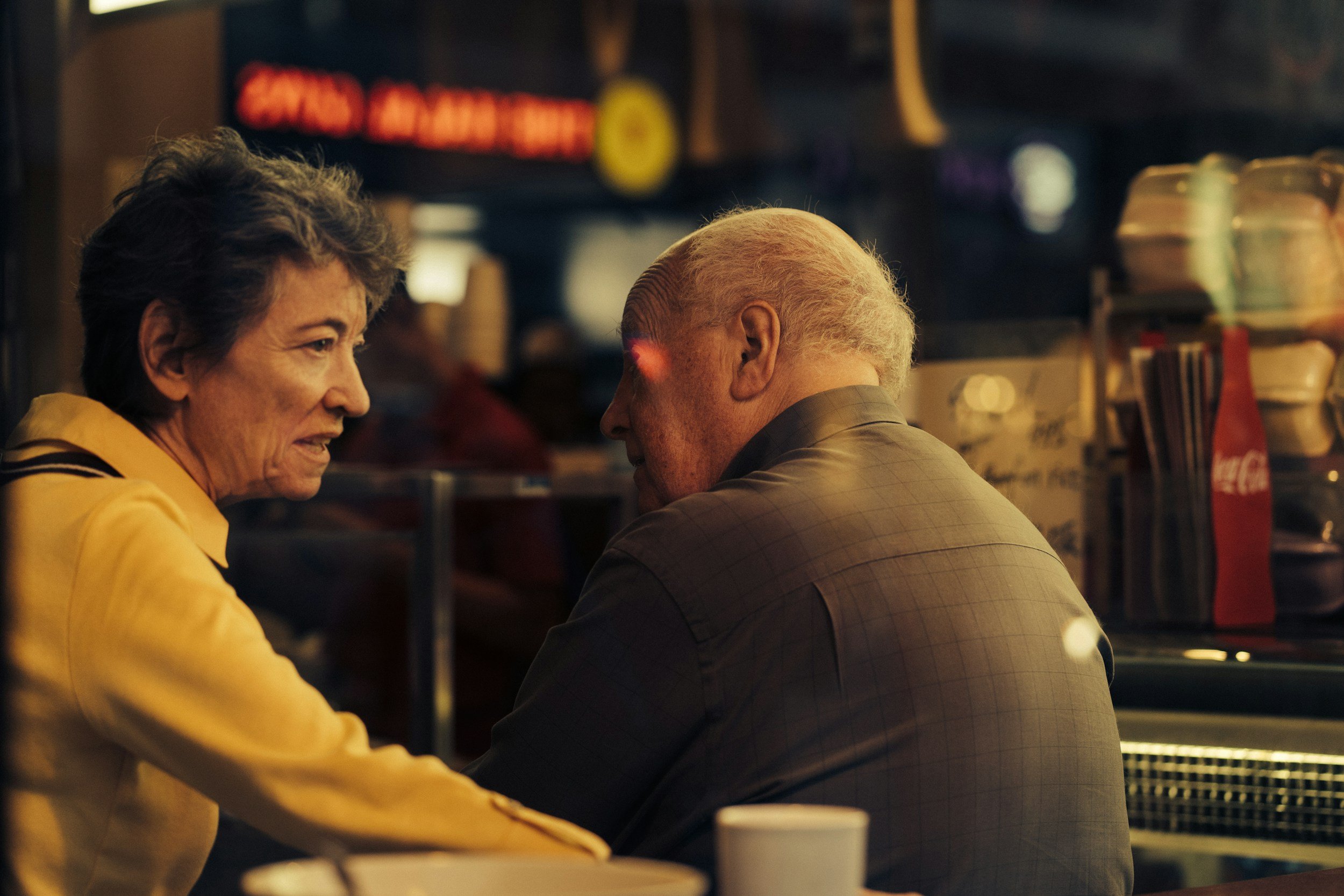 An elderly woman and man having a conversation at a bar or restaurant, with a dark, warm background and some illuminated signs.