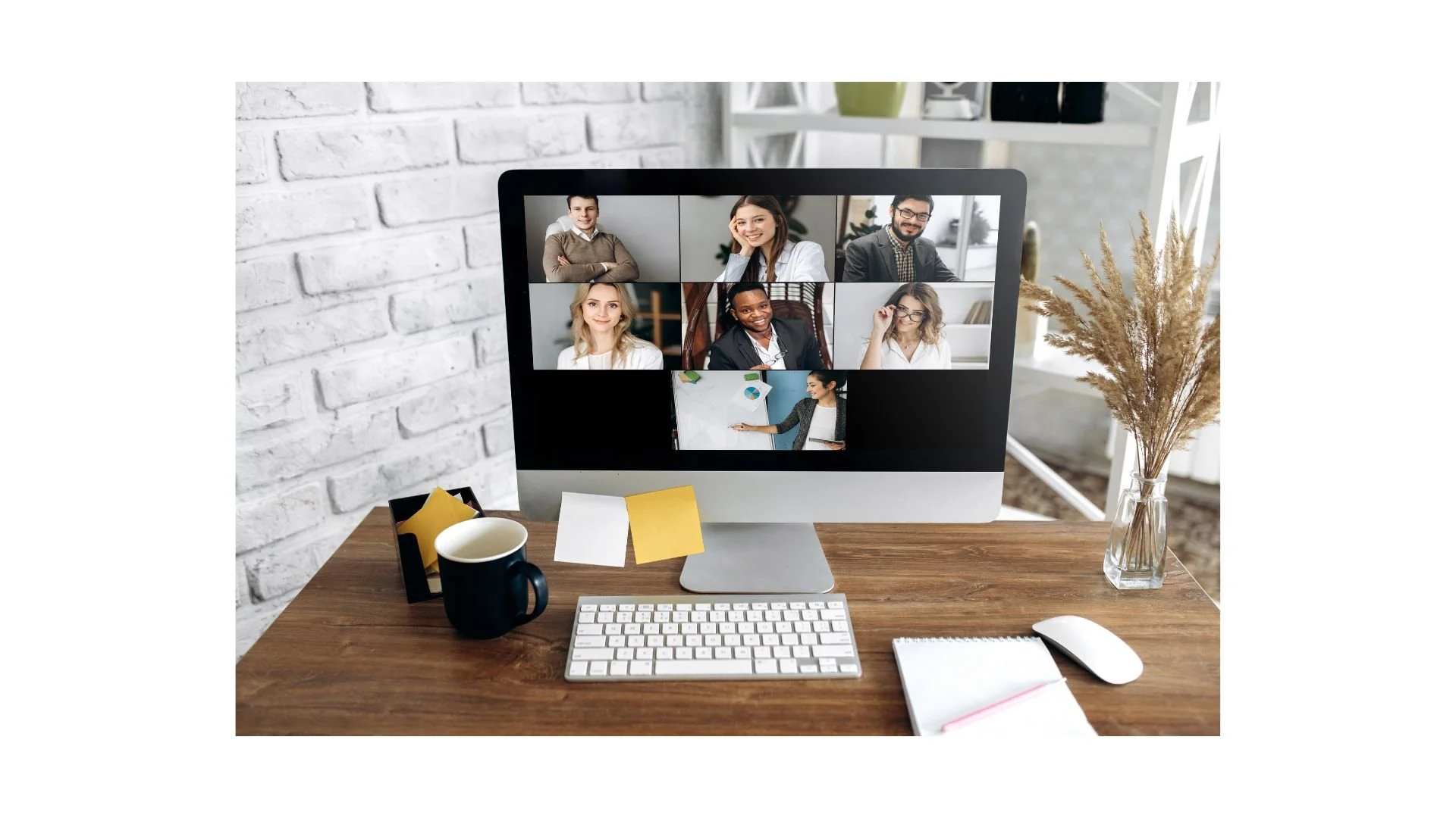 Computer monitor displaying a video conference with seven participants in a grid, on a wooden desk with a black mug, sticky notes, a keyboard, mouse, notepad, and a pen, with a vase of dried grasses on the right side and a white brick wall in the background.