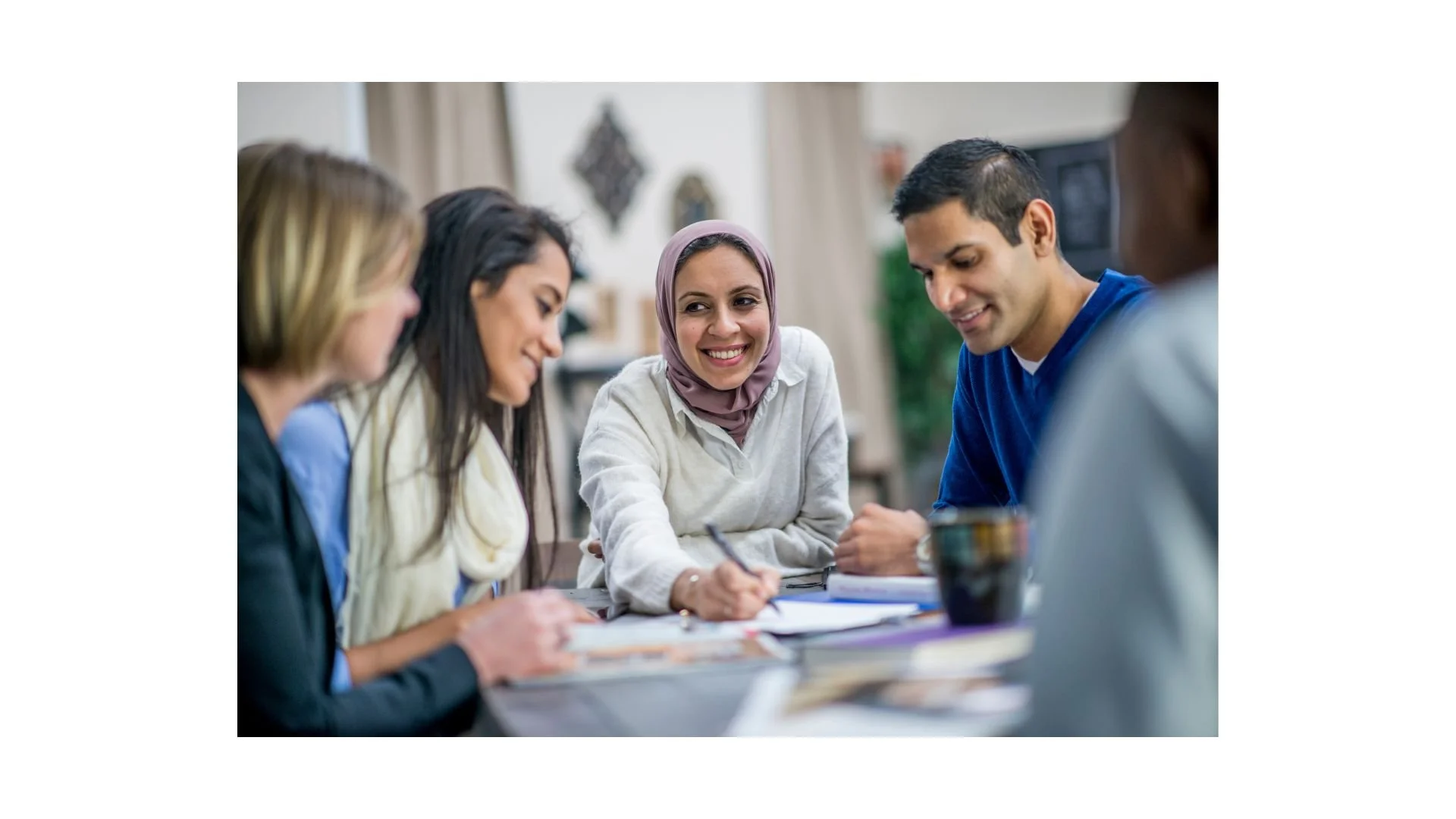 A diverse group of five young adults sitting around a table, engaged in a discussion or meeting, with one woman in a hijab smiling.
