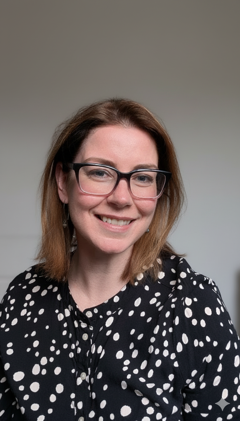 A woman with shoulder-length reddish-brown hair, wearing black-rimmed glasses, smiling, dressed in a black blouse with white polka dots.