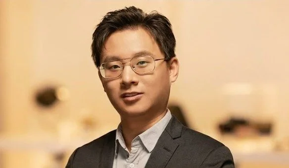 A young man with glasses, wearing a suit and white shirt, standing indoors with a blurred background.