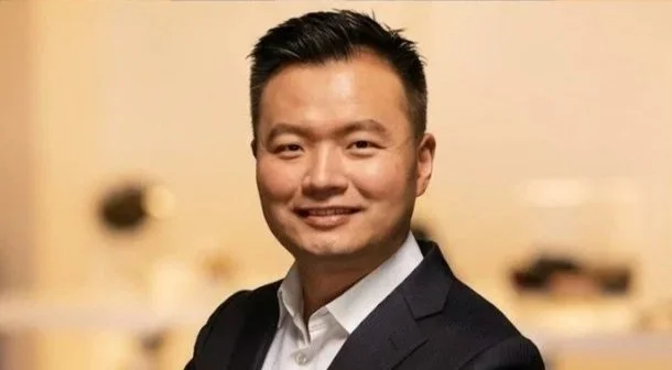 A man in a black suit smiling in front of a blurred background.