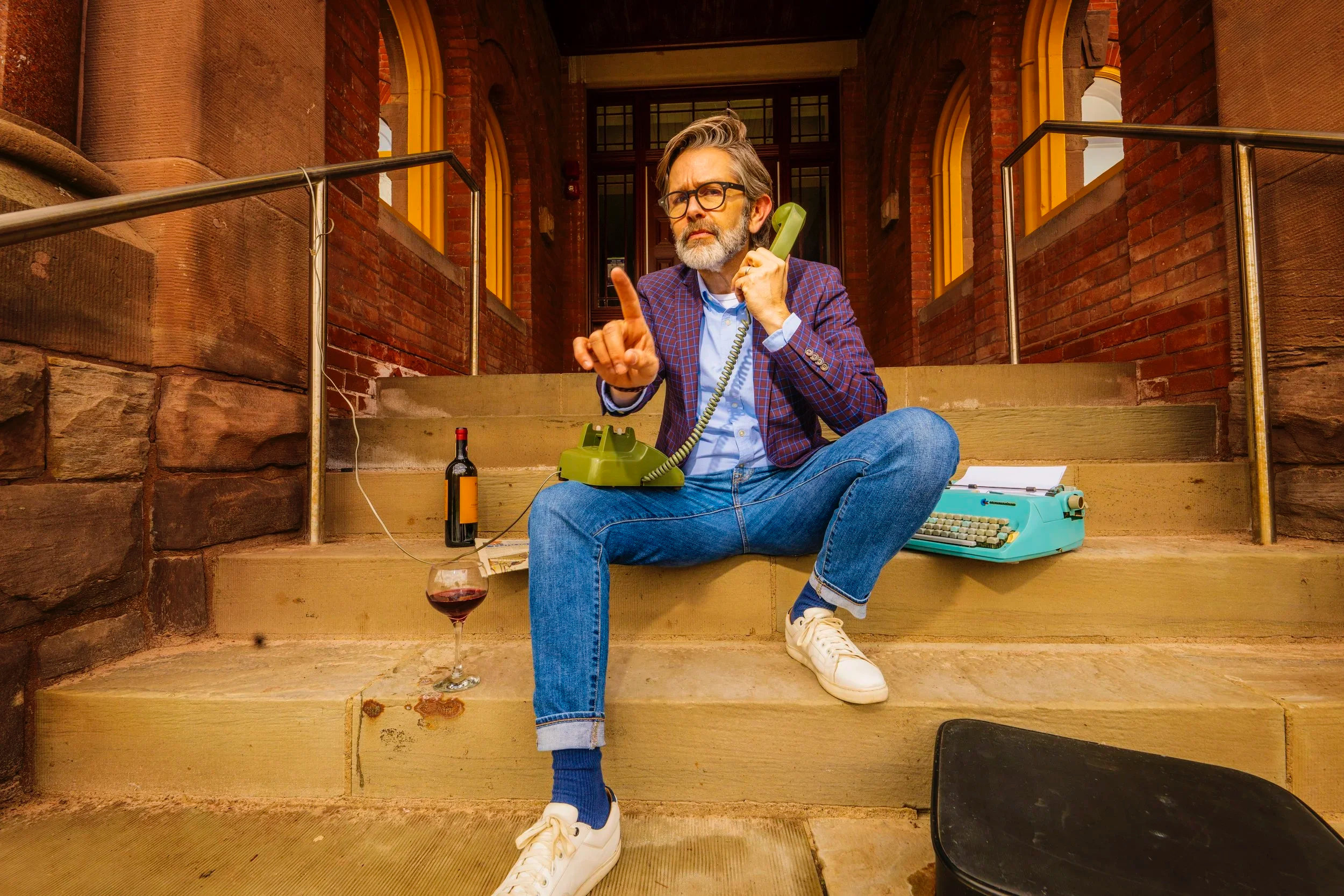 A man sitting on front steps of a brick building, multitasking with a vintage green telephone, a typewriter, and a wine glass with red wine nearby, wearing a checked blazer, jeans, and white sneakers, with a bottle of wine behind him.