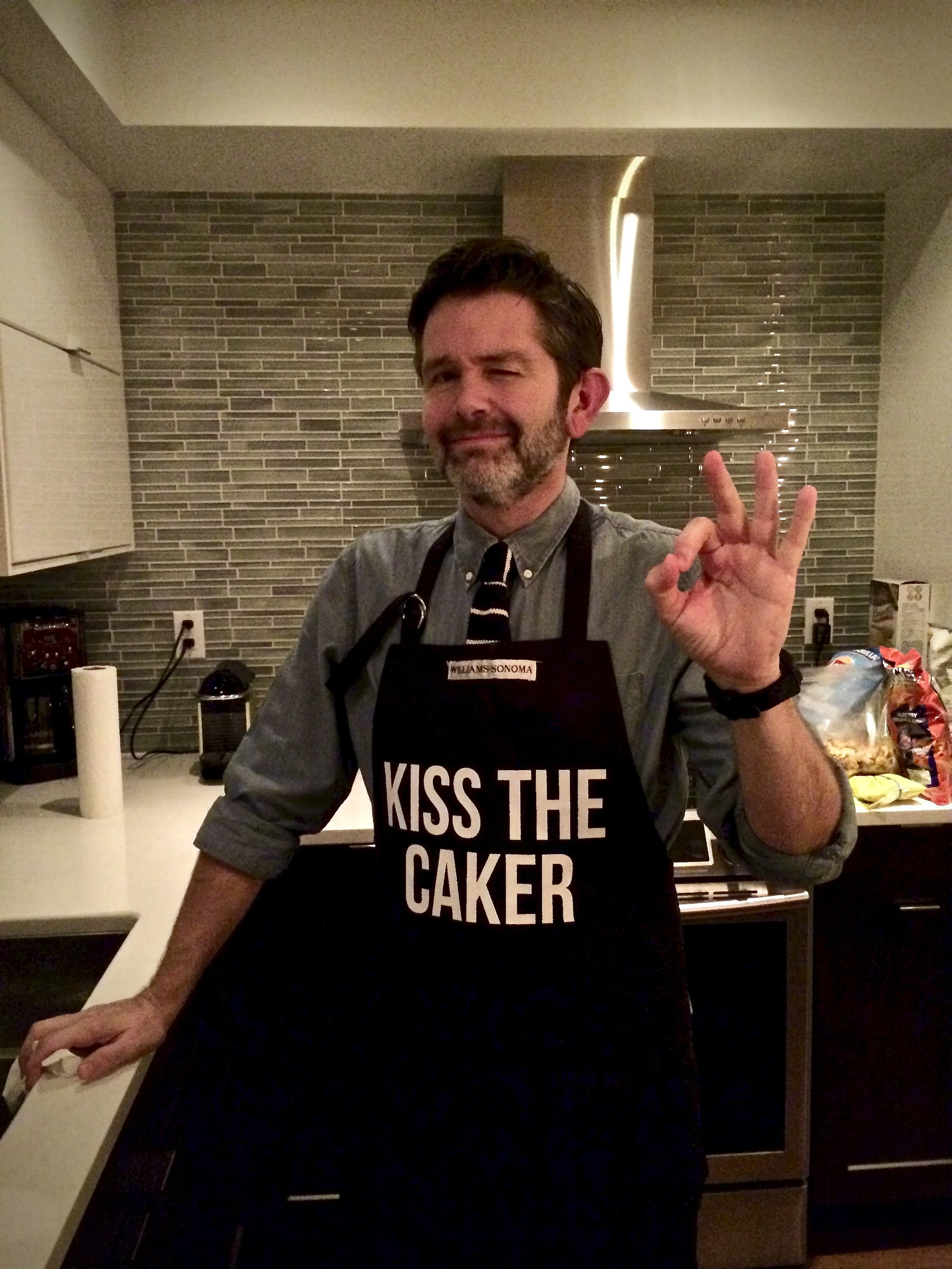 Man wearing an apron that says 'Kiss the Caker' in a home kitchen, making an OK hand gesture.