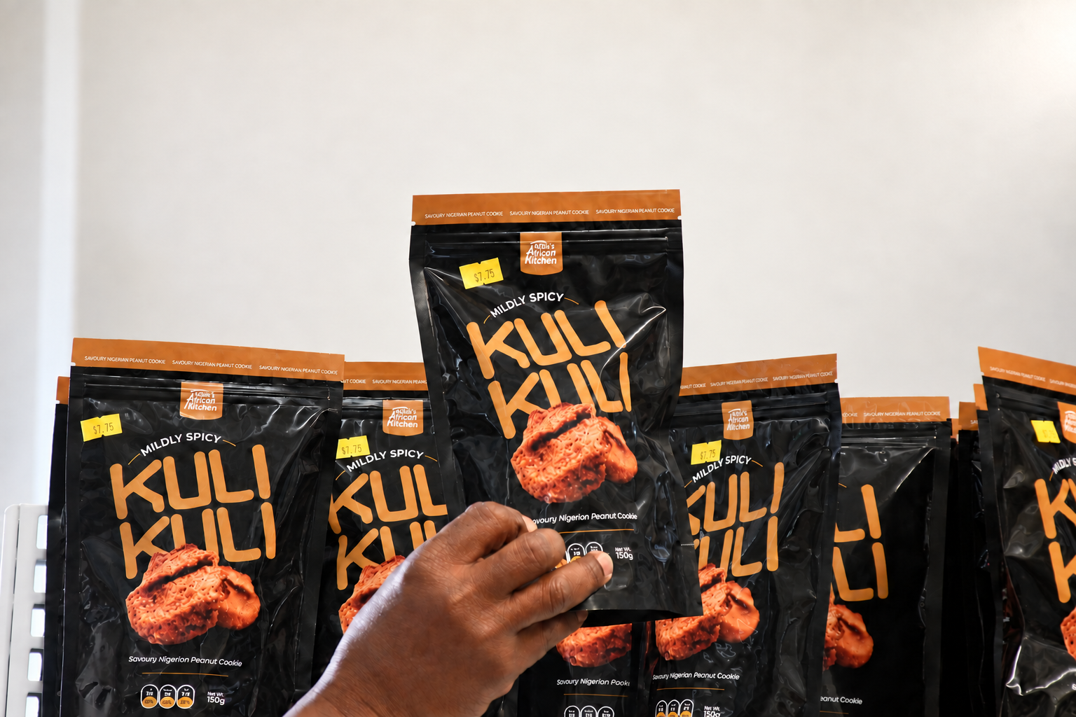 Person holding a black bag of mildly spicy Kuli Kuli Nigerian peanut cookies on a store shelf, with similar bags behind.