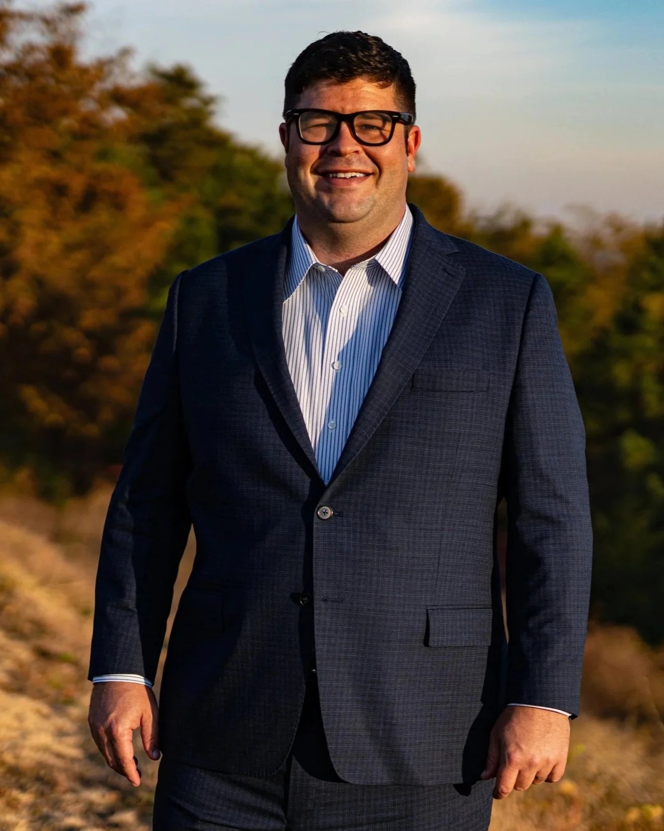 A man wearing glasses and a dark blue suit standing outdoors during sunset with trees in the background.