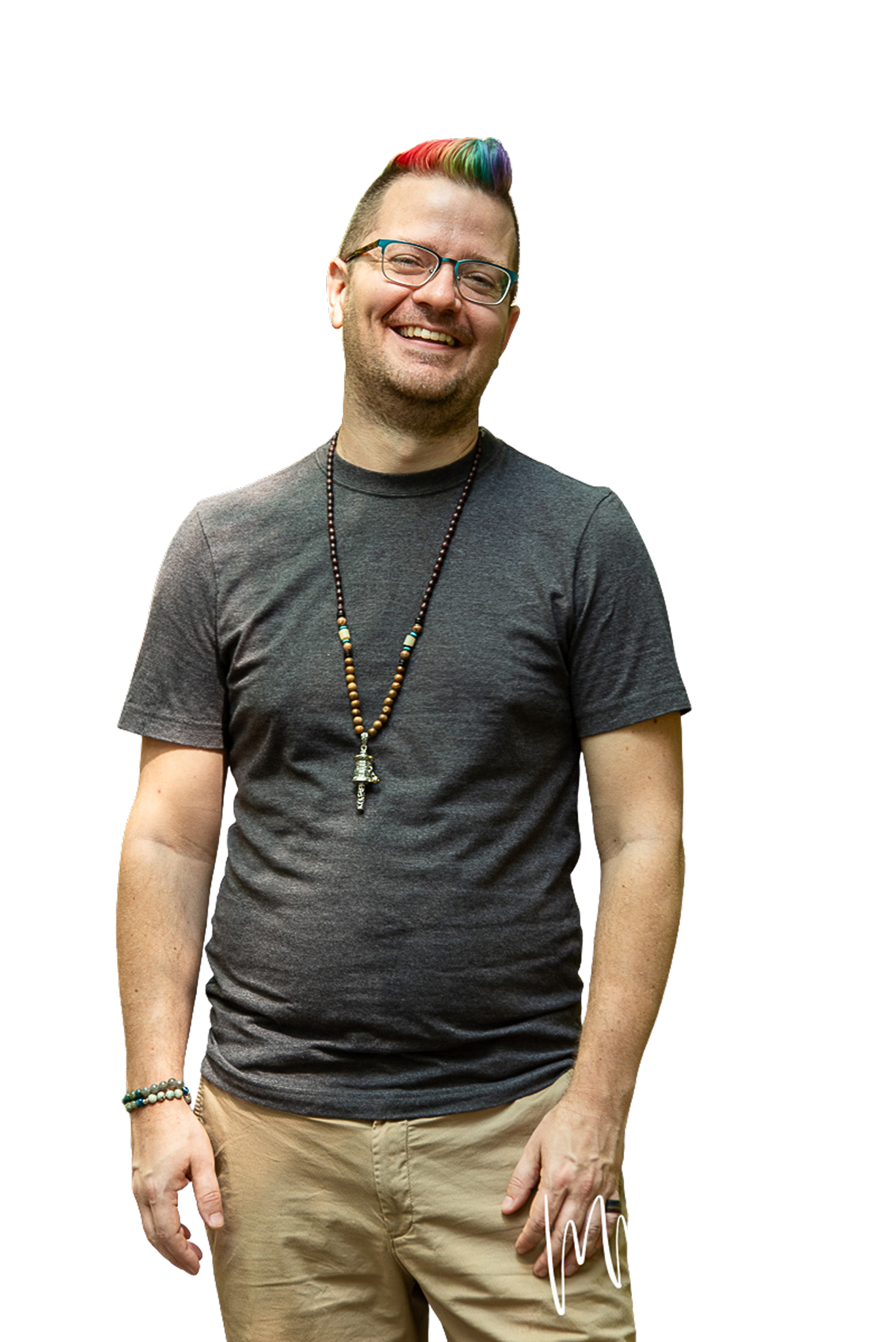 A smiling man with glasses, colorful hair, and a beard, wearing a gray t-shirt, tan pants, and beaded accessories, standing outdoors.