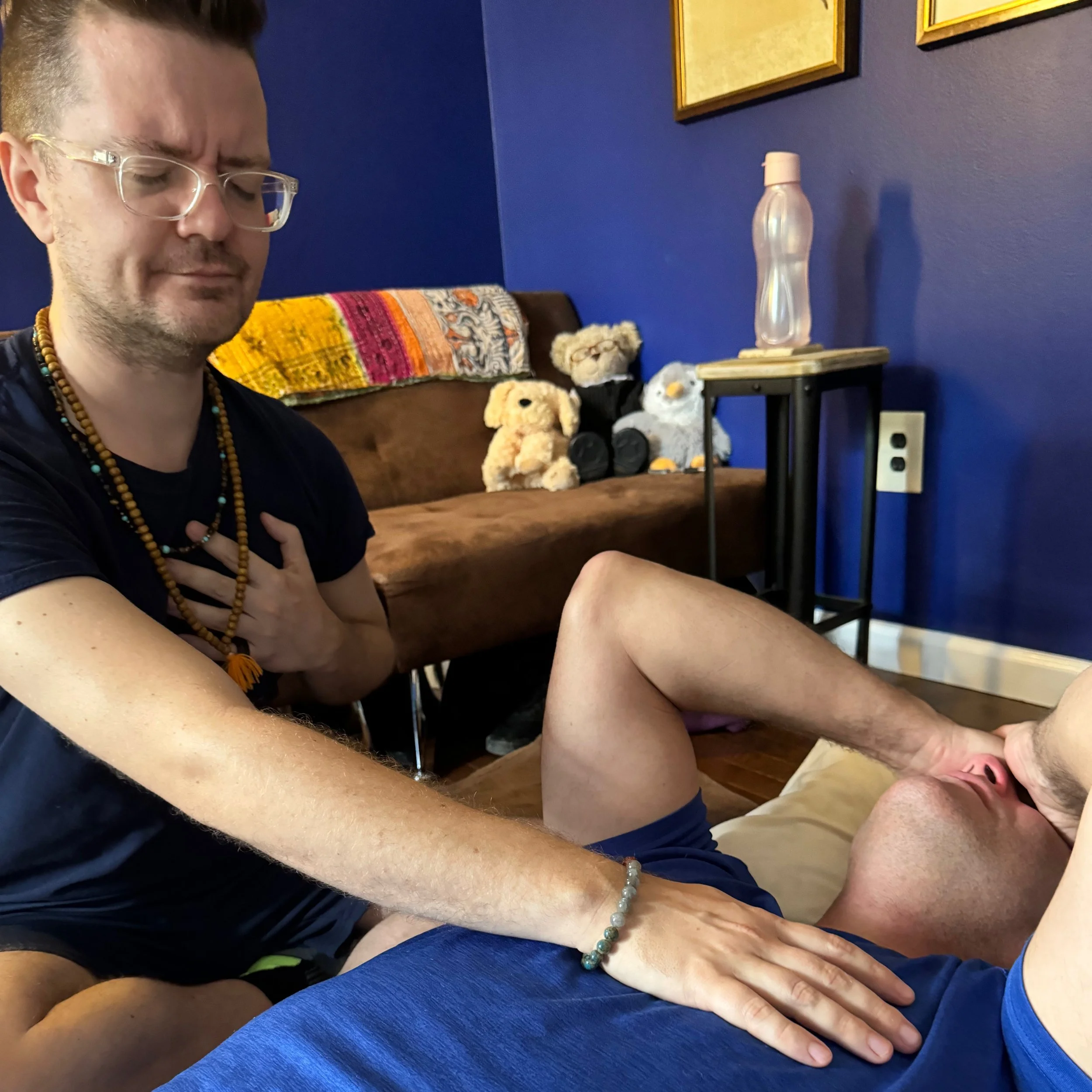 A person with glasses and beaded necklaces holding their hand on another person's chest while they are lying down on a bed. The person on the bed has their hand on their face and is wearing a blue shirt. In the background, there is a brown couch with several stuffed animals on it, and a blue wall with framed pictures.