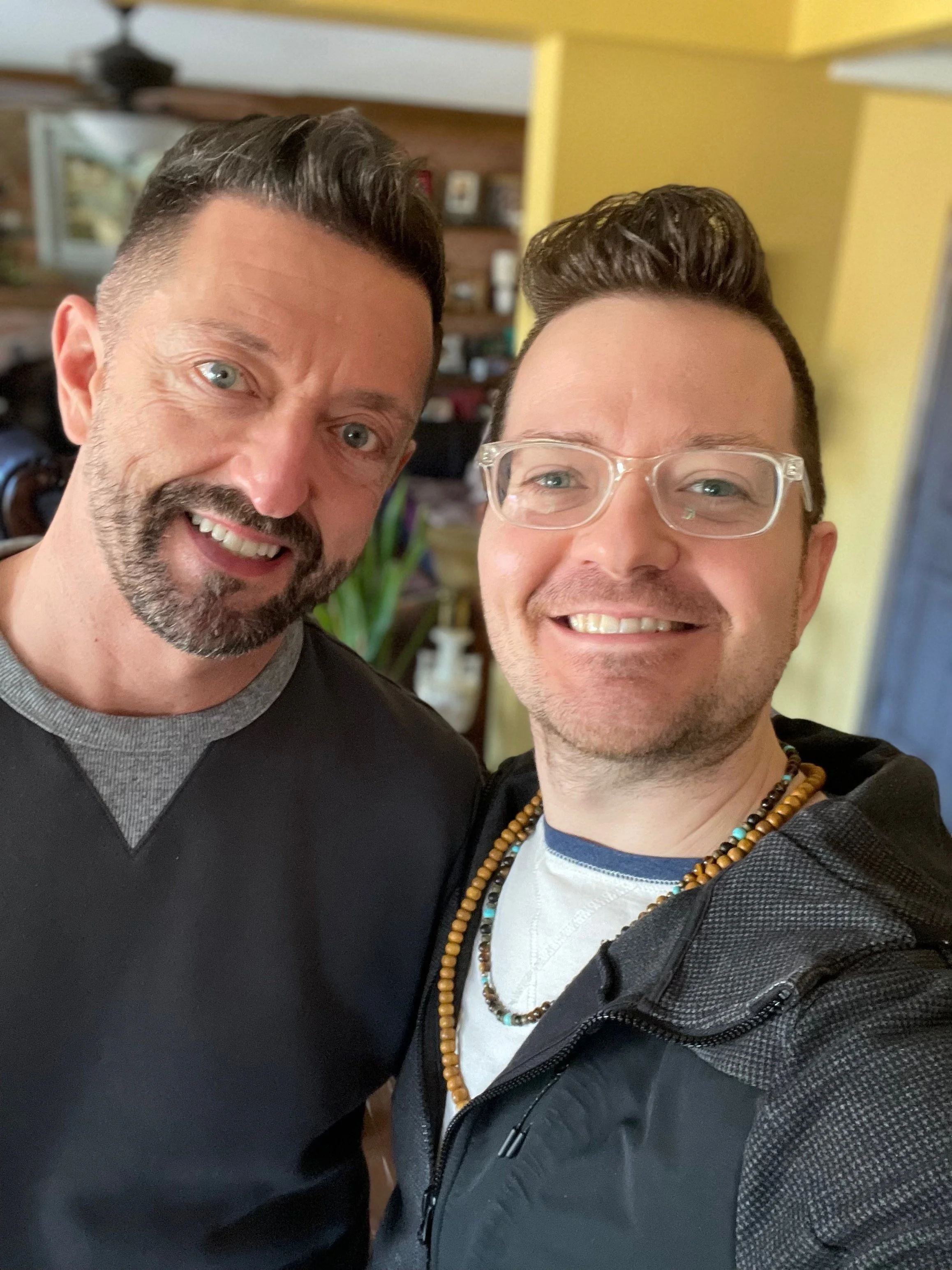 Two men smiling for a selfie indoors, with bookshelf and plants in the background.