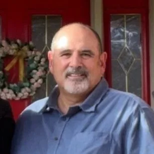 A man with short hair, a beard, wearing a blue shirt, standing outside in front of a window and a floral wreath.