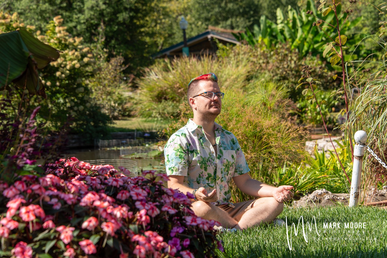 A man with rainbow-colored hair and glasses sitting cross-legged on grass, meditating in a lush garden with flowers, trees, and a small water feature.
