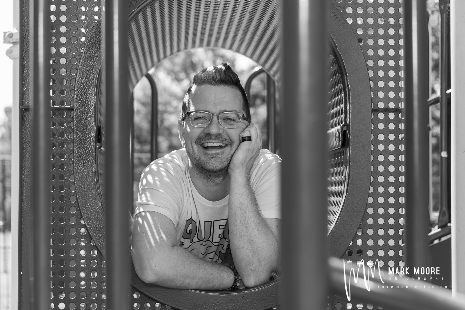Man with glasses smiling, lying on playground equipment, resting his head on his hand, with sunlight and trees in background.