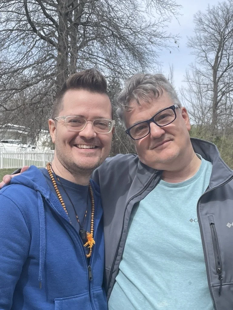 Two men smiling in an outdoor setting with trees and a white fence in the background. They are close together, one wearing a blue hoodie and the other wearing a light blue T-shirt and gray jacket.