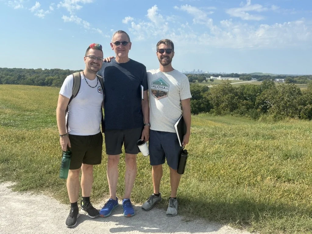 Three men standing outdoors on a sunny day with a grassy field and trees in the background. They are smiling and dressed casually with water bottles and a tablet.