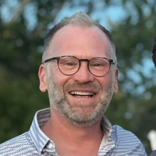 A man with glasses and a beard smiling outdoors with blurred trees in the background.