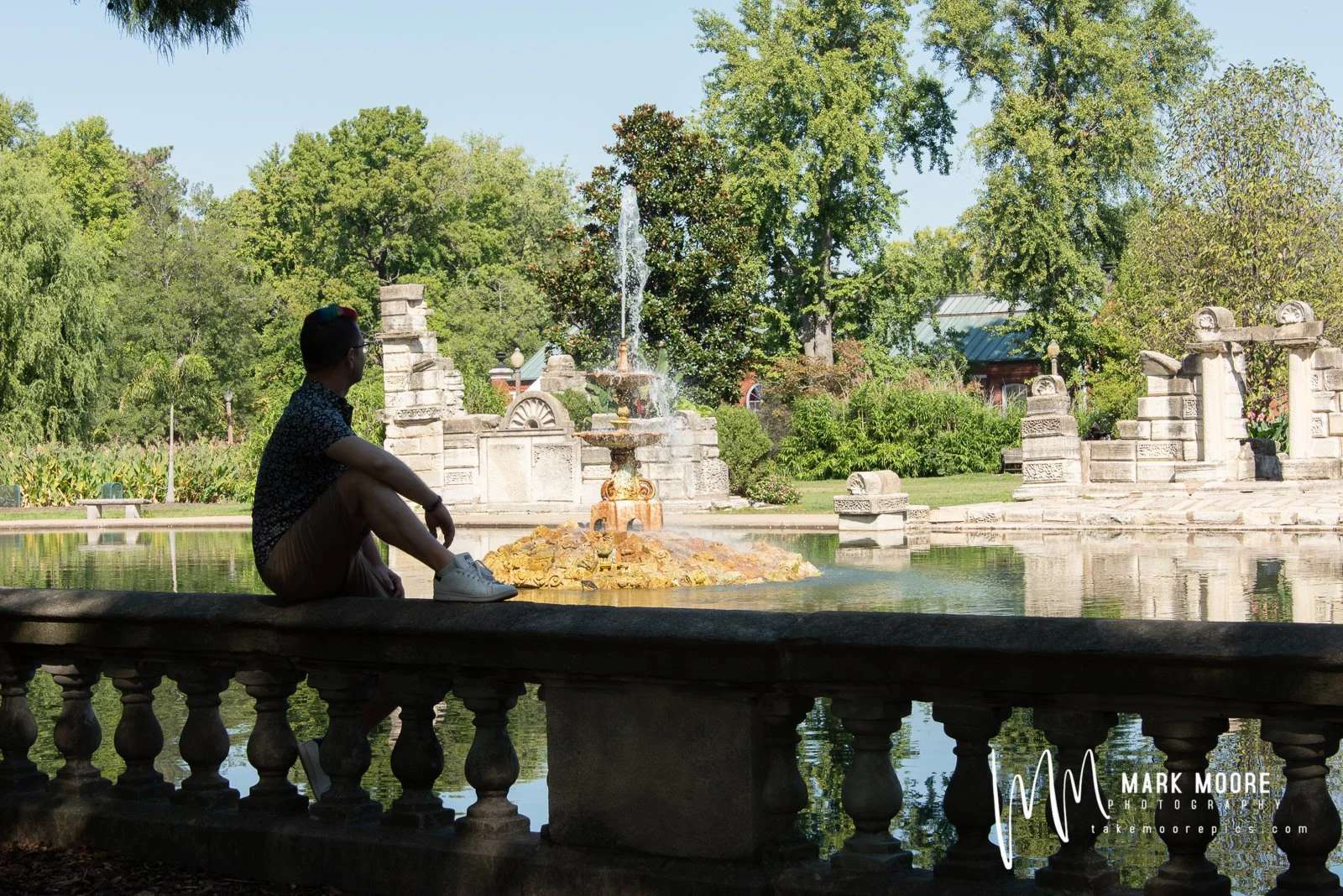 A person sitting on a stone barrier by a pond with a fountain, surrounded by green trees and stone arches on a sunny day.