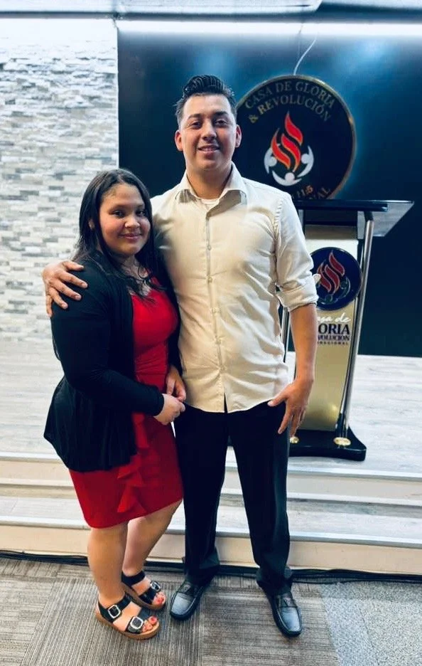 A young man in a white shirt is stadning with wife and a logo behind him with white and red flames