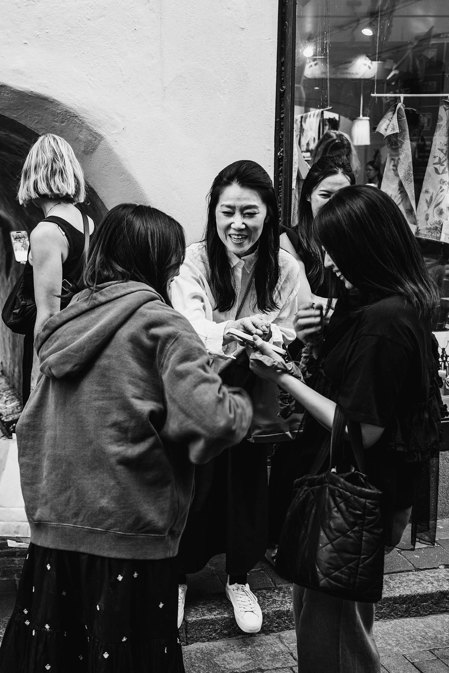 Black and white photo of a group of young women gathered on a street, interacting and looking at a phone.