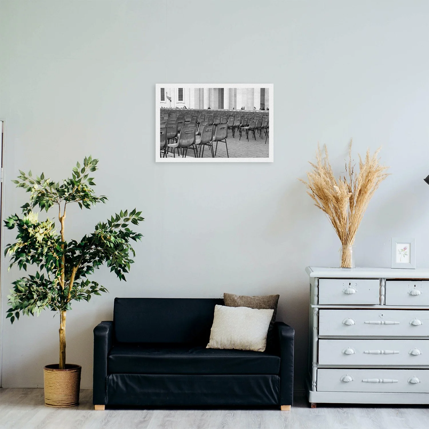 A room interior featuring a black sofa, potted tree, light gray dresser, and framed black and white photo of empty rows of chairs outside a classical building.