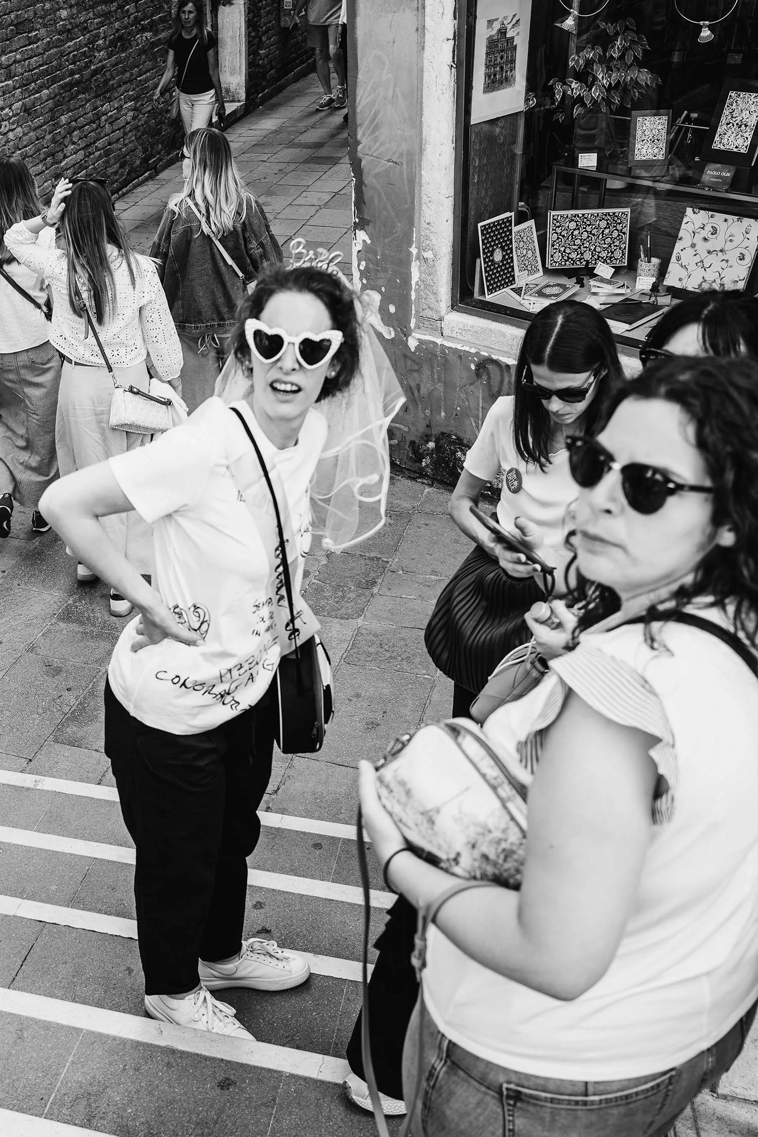 Group of women standing and interacting on a narrow street near a wall and shop window displaying artwork, in black and white.