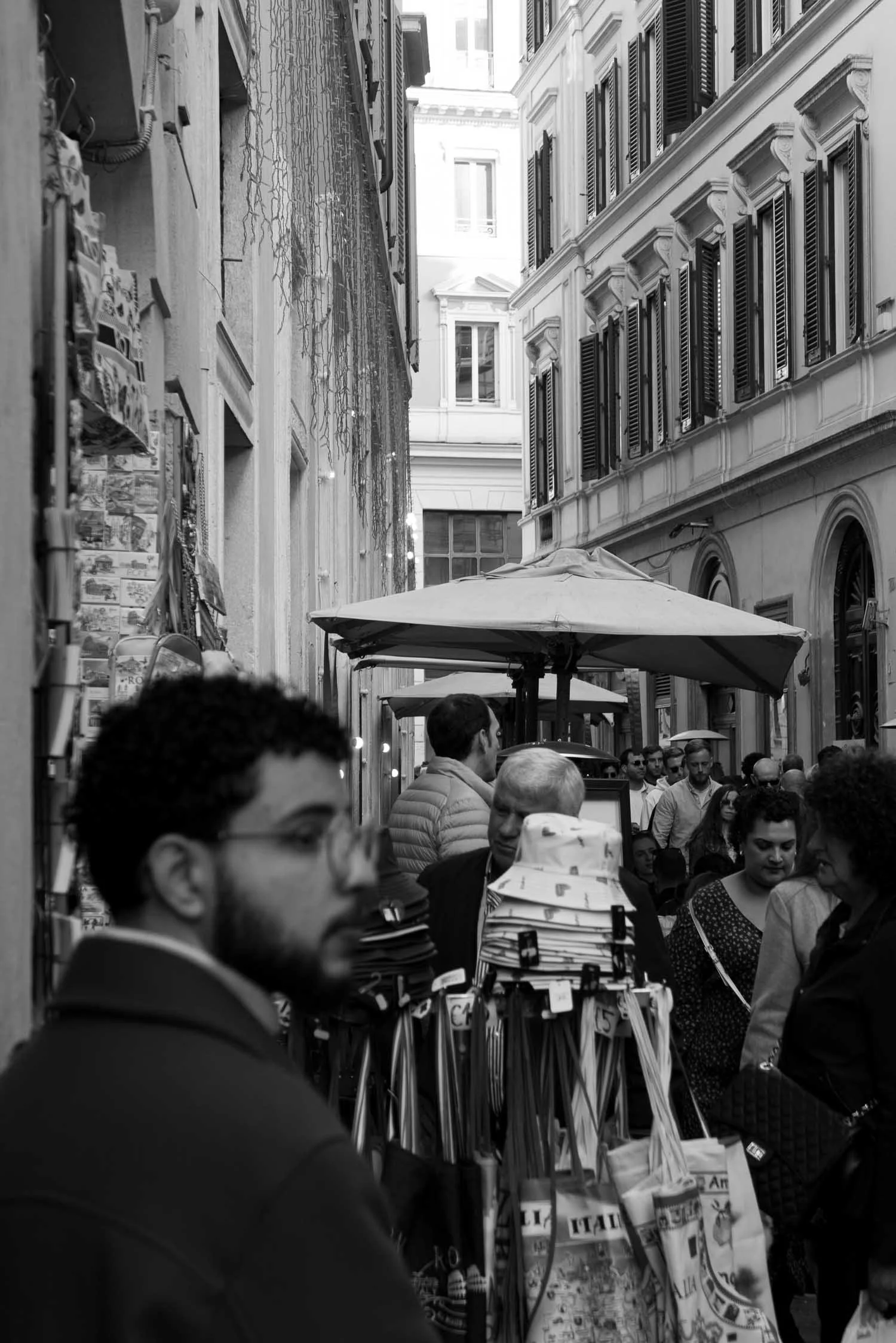 Crowded, narrow European street with shop displays featuring hats, bags, and souvenirs under umbrellas, lined with historic buildings.