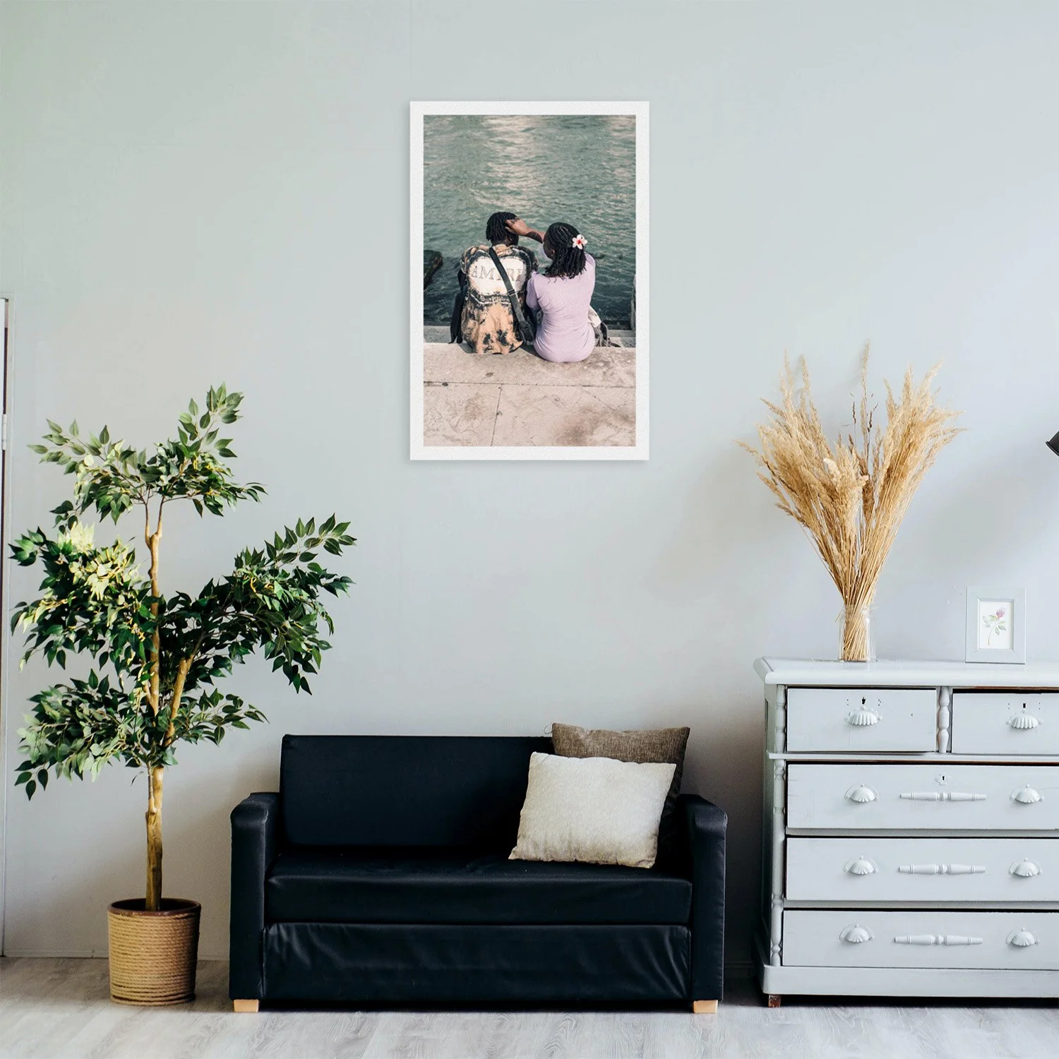 Two people sit on stone steps facing a body of water, with a framed photograph of them hanging on the wall above a black sofa.