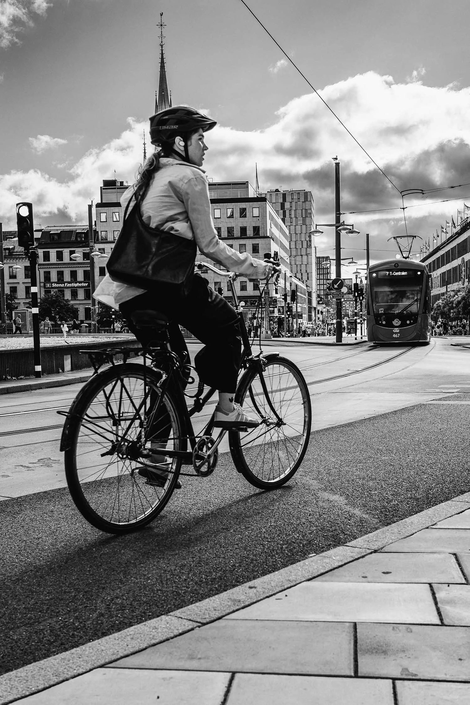 Person wearing a helmet rides a bicycle near a tram in an urban cityscape with buildings and cloudy sky in black and white.