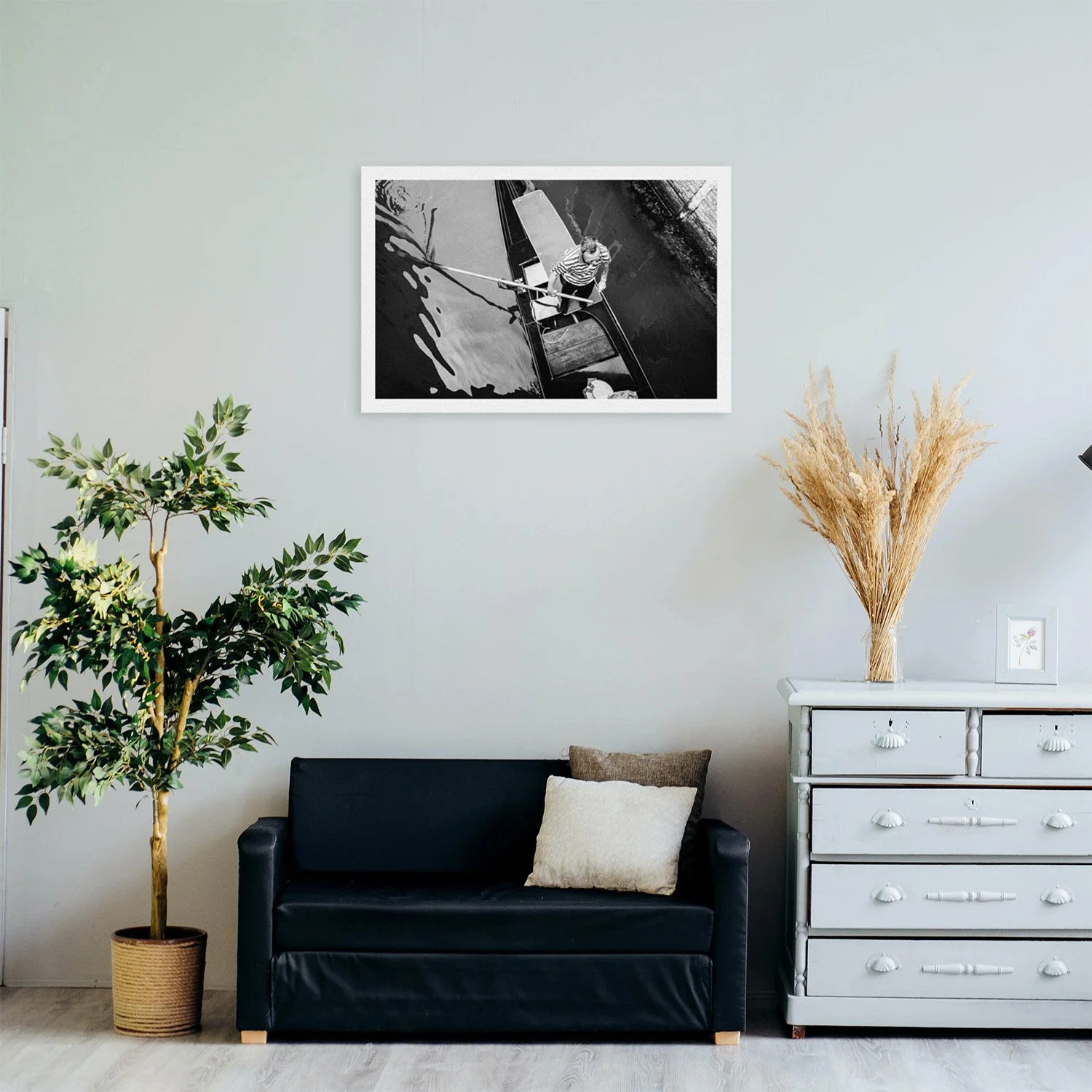 Bright living room with black sofa, potted tree, white dresser, dried grasses, and framed black-and-white boat photograph.