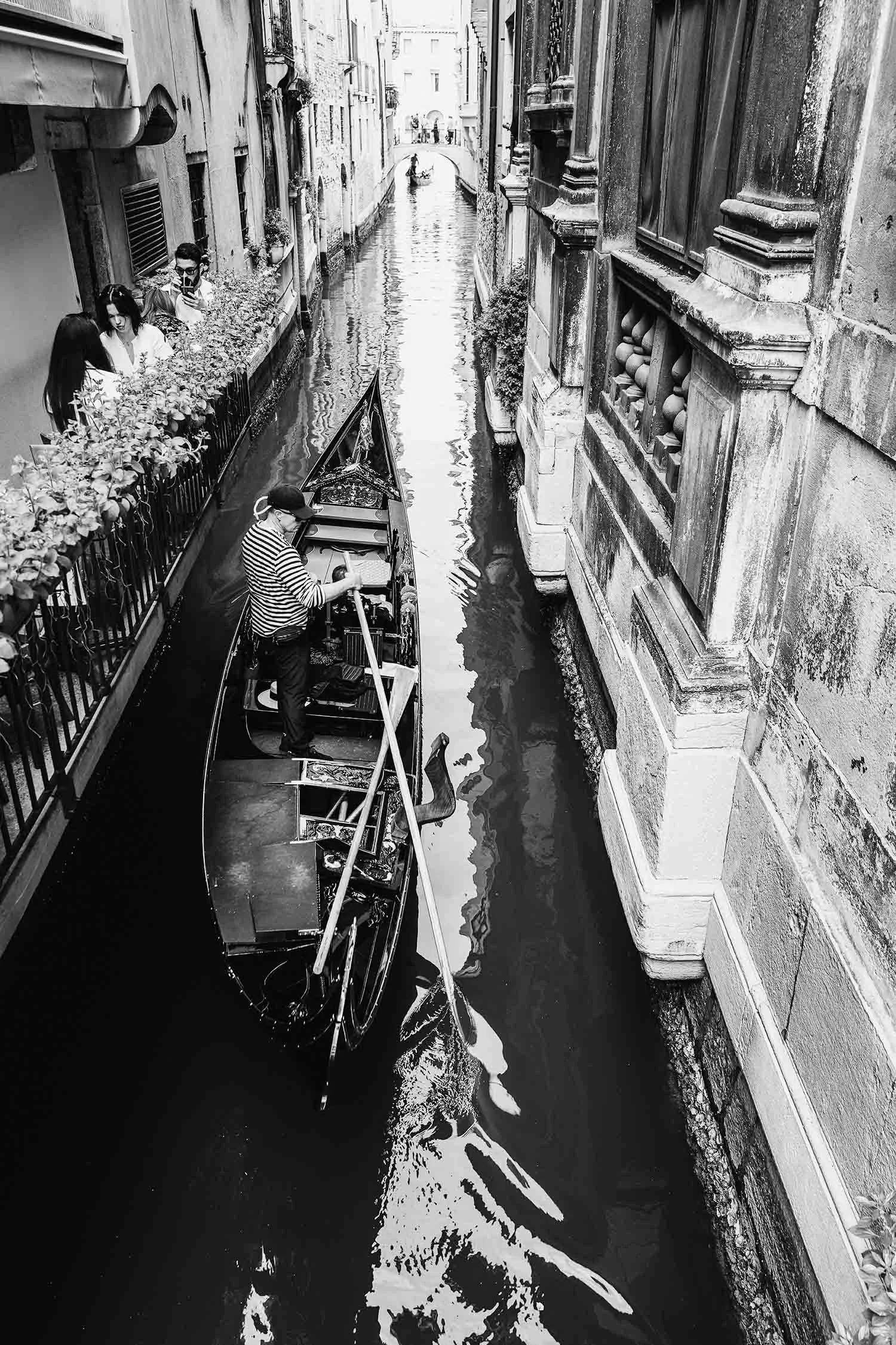 Black and white photo of a gondolier steering a gondola through a narrow Venetian canal with historic buildings on each side.