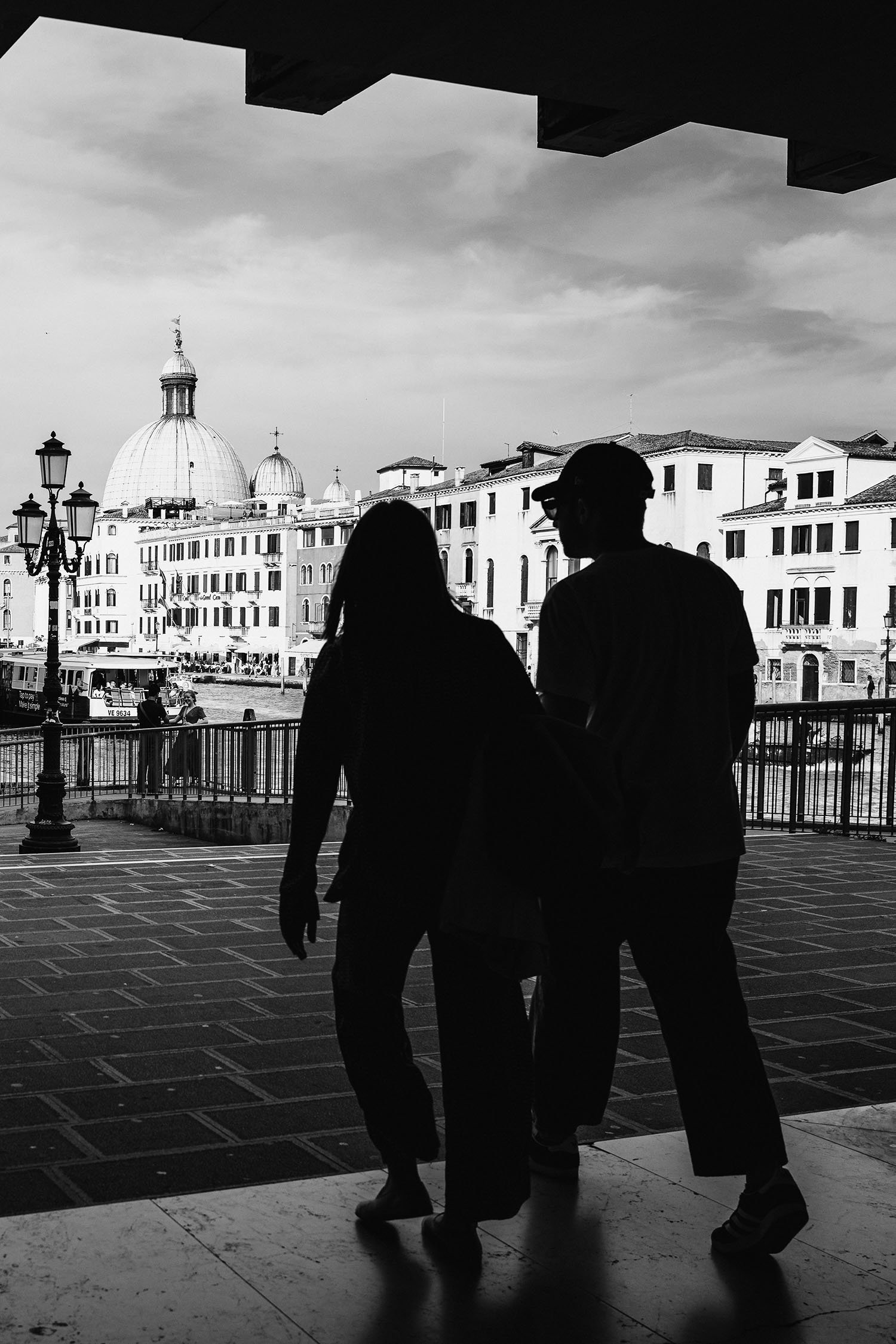 Silhouetted couple walking hand-in-hand, framed by an archway, with Venetian architecture and a dome in the background.
