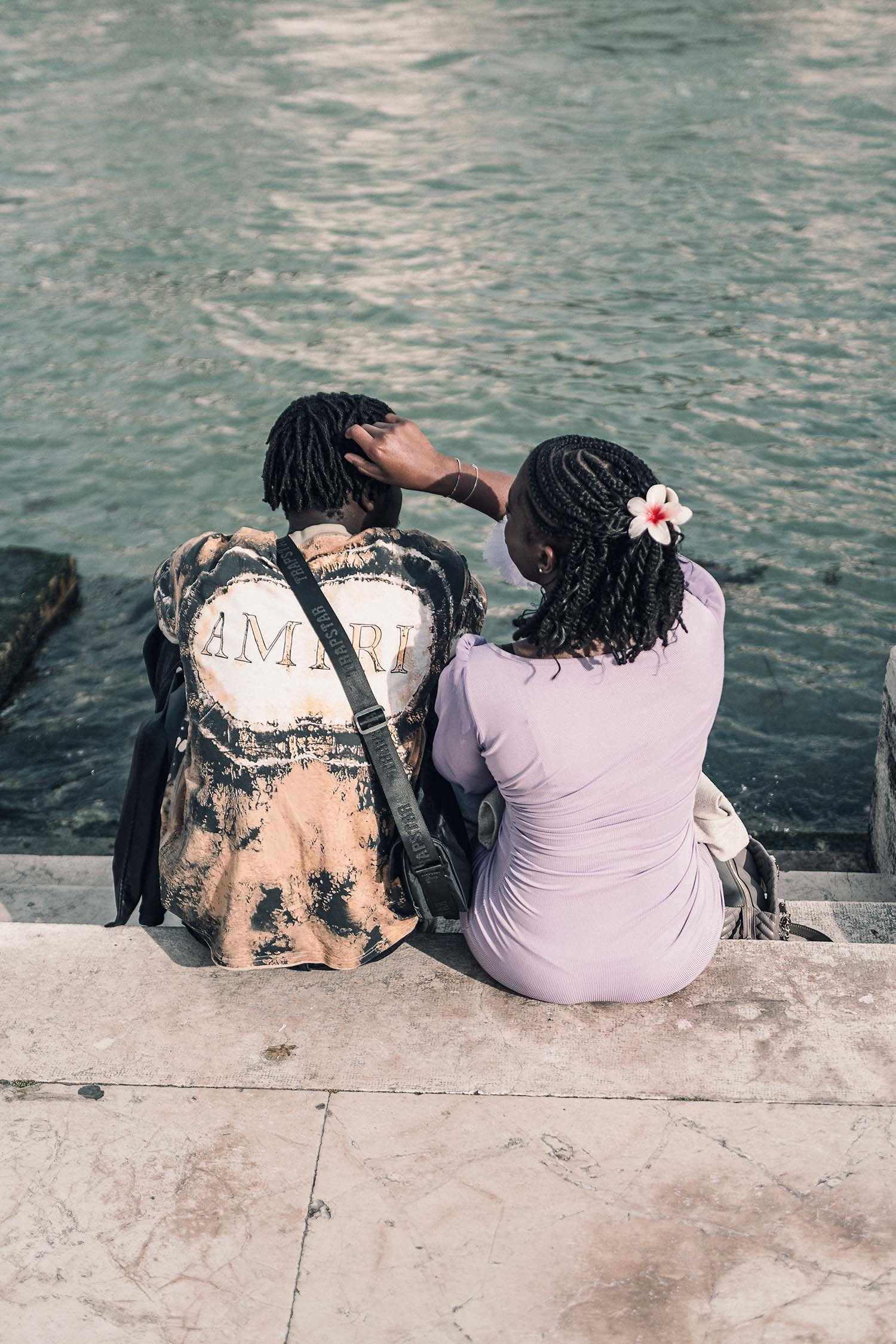 A couple sits on a ledge by the water, the woman gently touching the man’s hair, both in casual, stylish outfits.