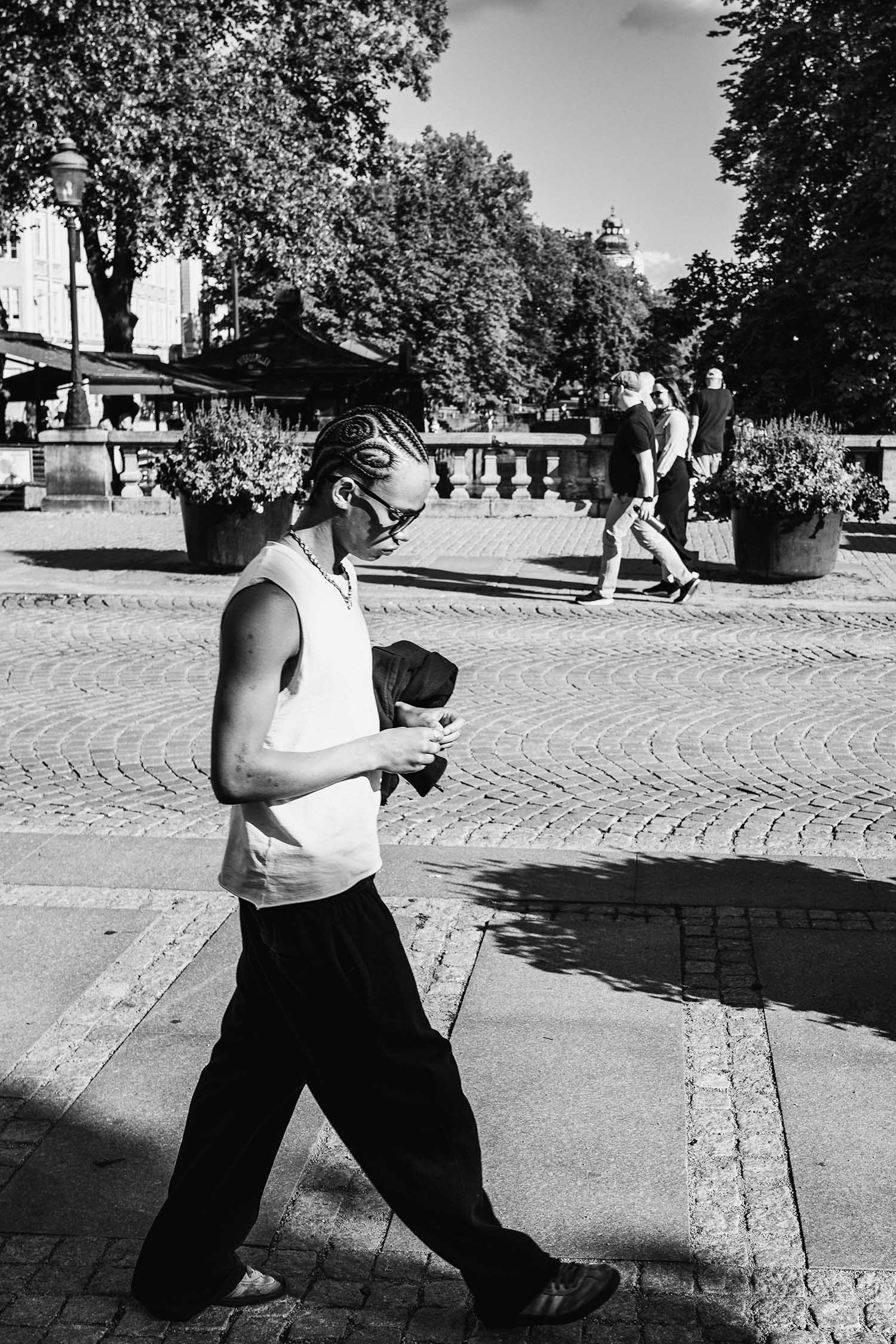 Black and white photo of a person with braided hair walking on a sunny cobblestone street, wearing sunglasses and a sleeveless shirt.